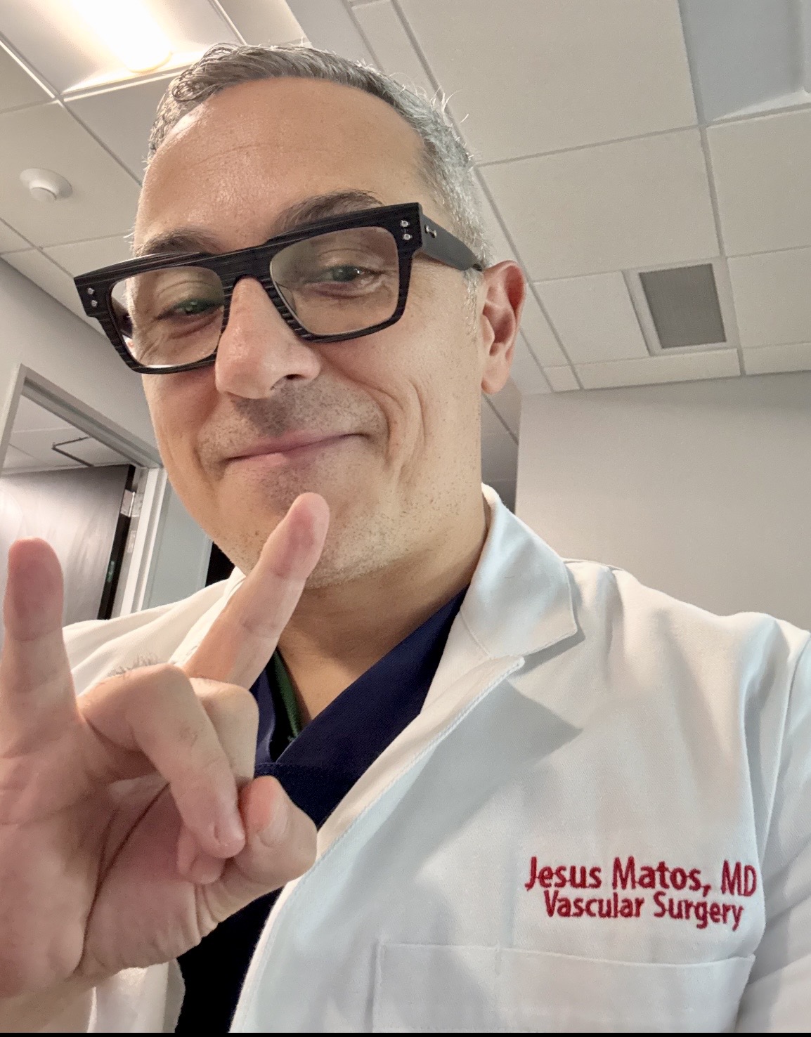 Man in white coat with glasses making a peace sign, smiling, in a clinical setting.