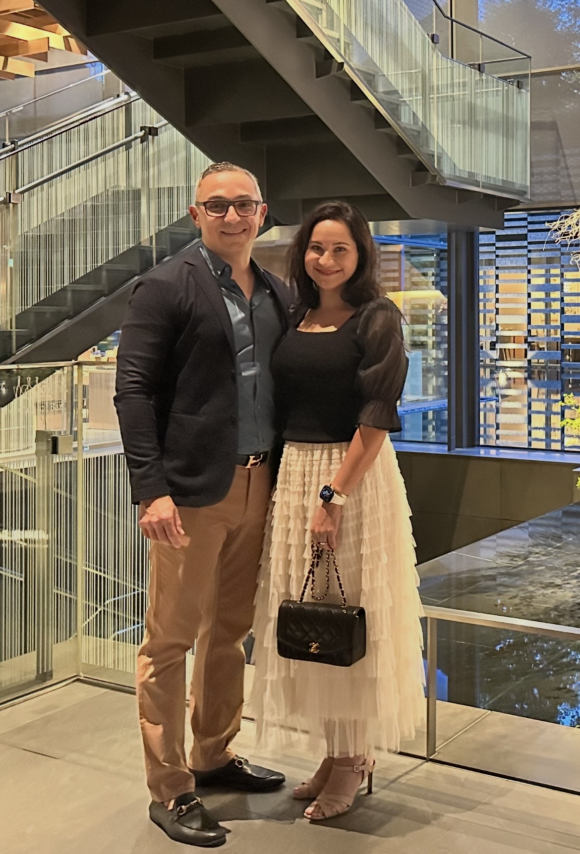 A man and woman stand together indoors, smiling, with a modern staircase and cityscape visible behind them.