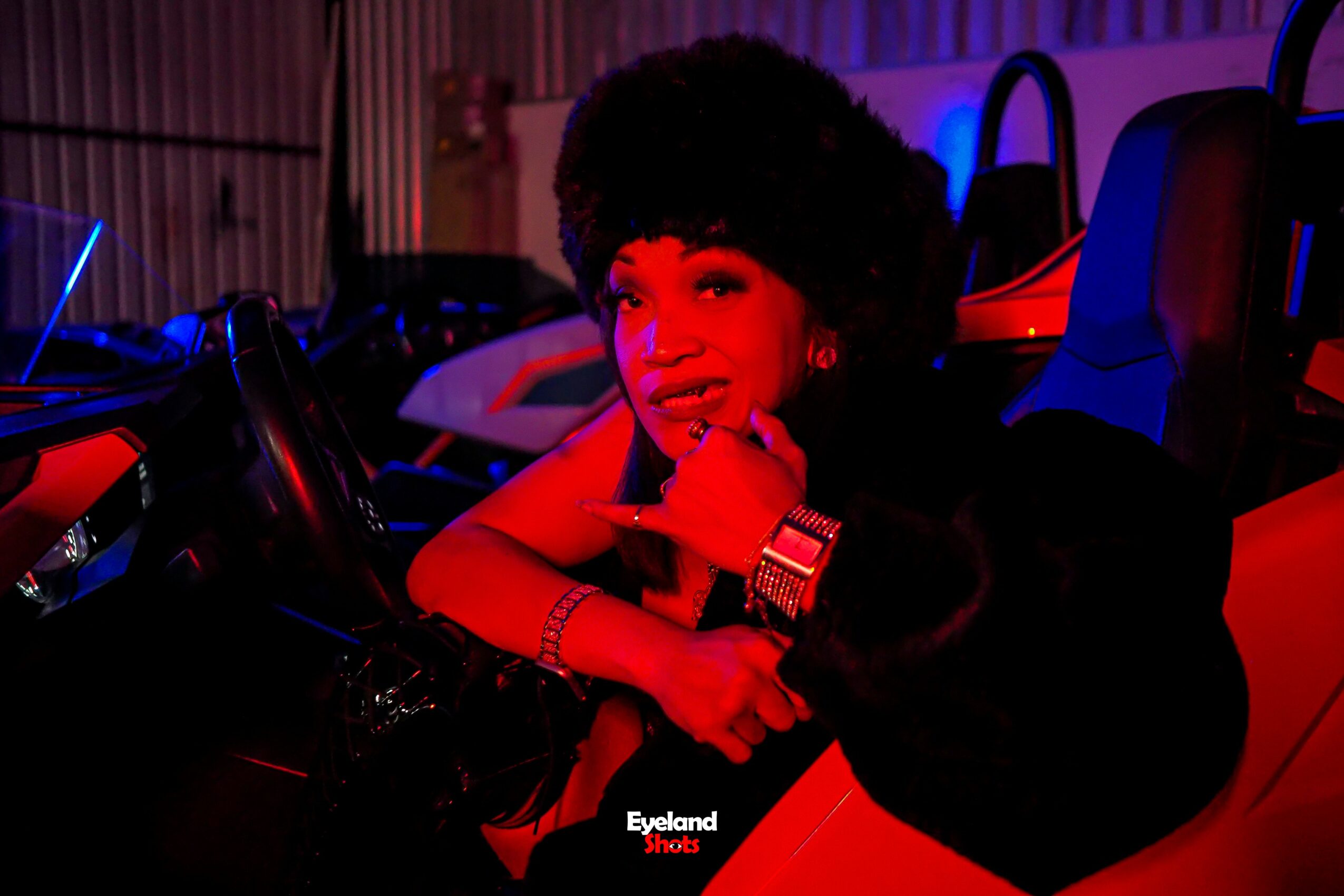 Person with large curly hair sitting in a red and black vehicle, smiling, with colorful lighting in the background.