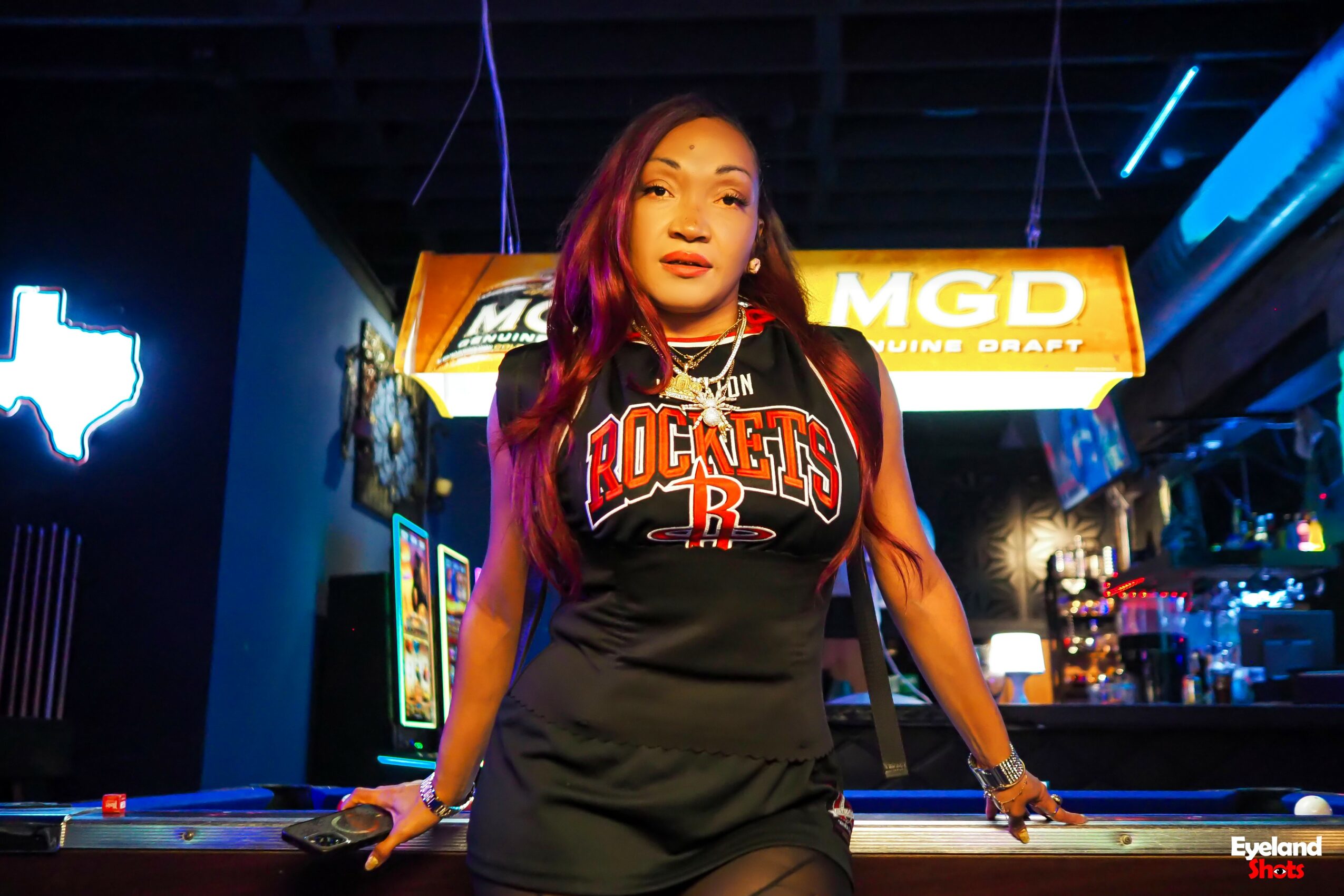 Woman with long hair wearing a Houston Rockets jersey, standing in a bar or arcade setting with neon signs.