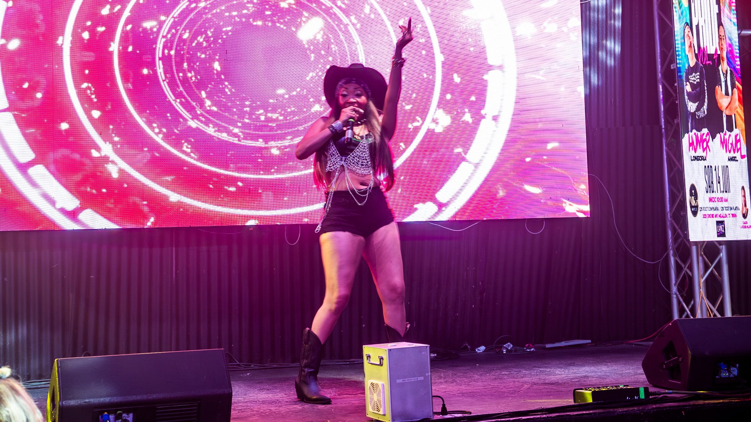 Performer on stage singing into microphone with pink circular digital background, wearing black hat, shorts, and boots.