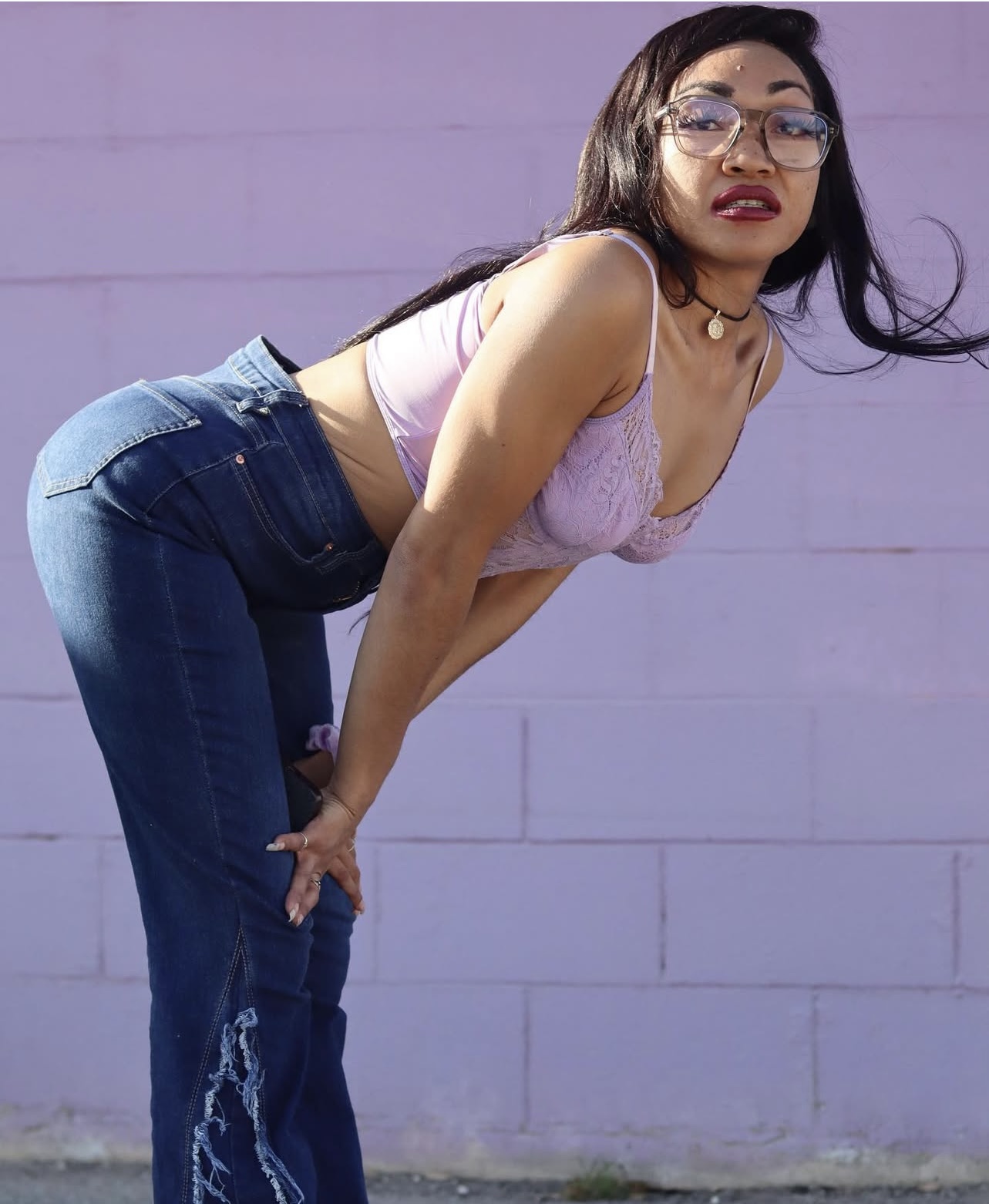 Woman with glasses leaning forward, wearing a pink top and jeans, standing against a purple wall.
