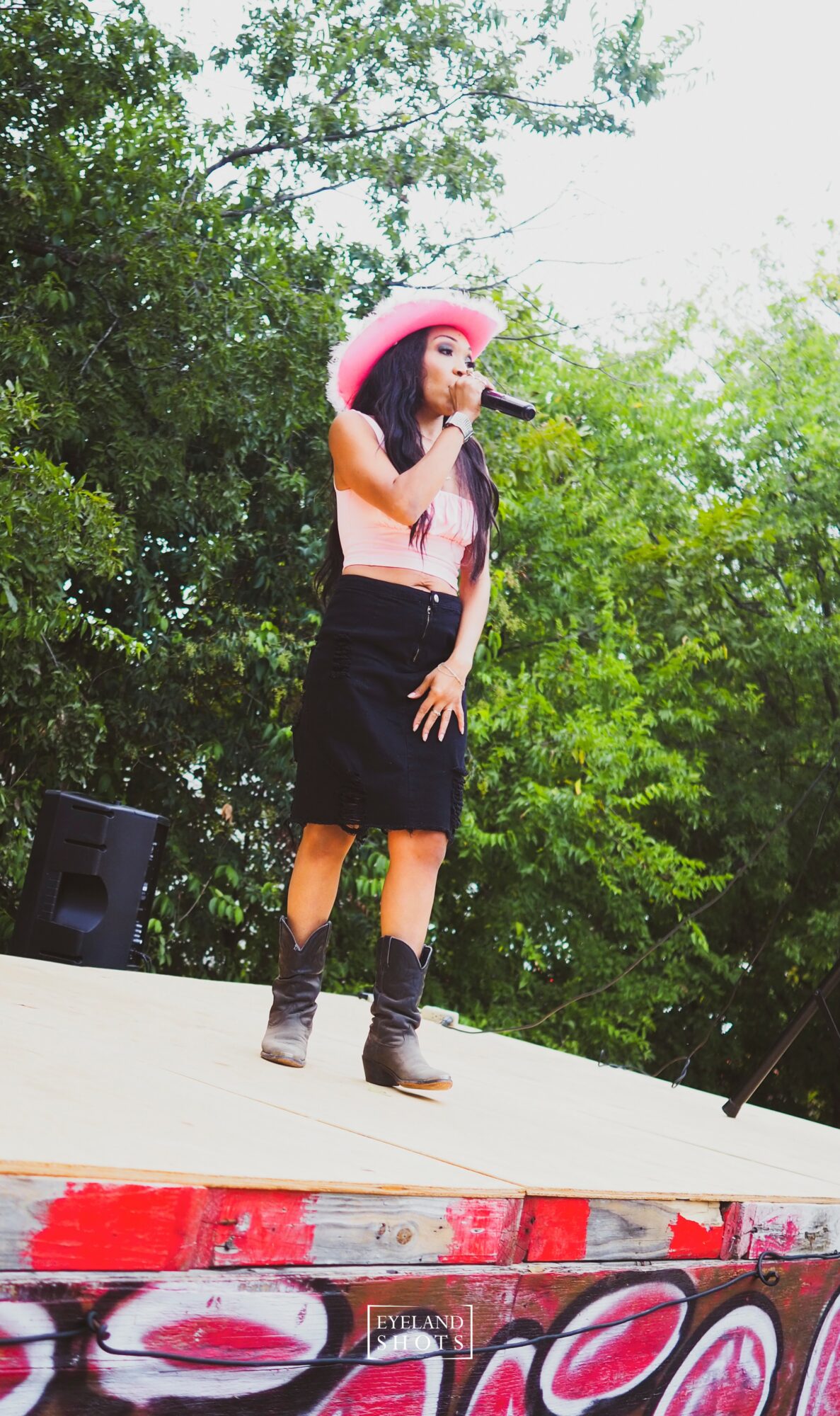 Young woman wearing a pink hat, black skirt, and boots singing into a microphone on an outdoor stage with trees in the background.