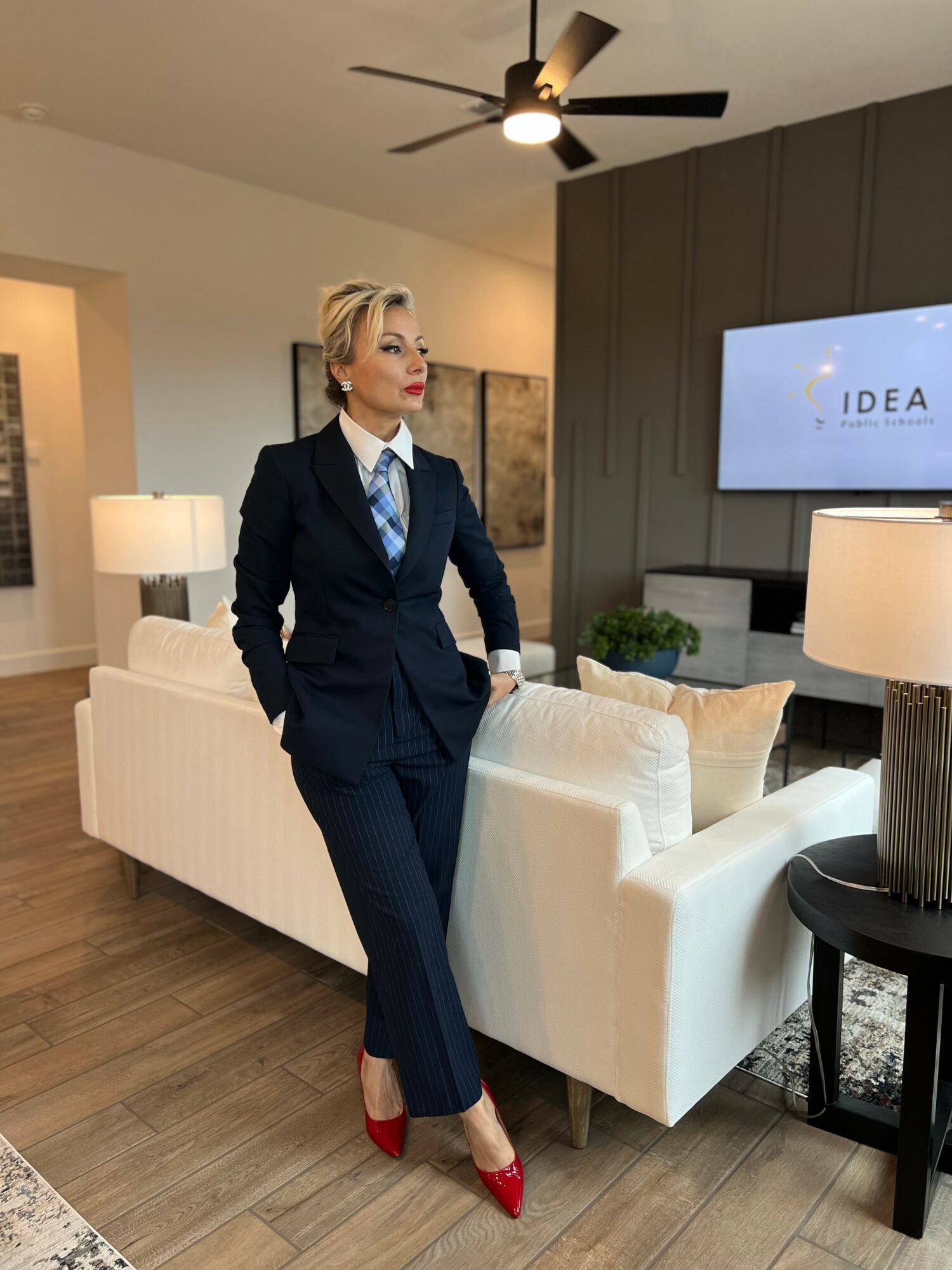 Woman in a navy suit with a blue tie and red heels standing in a modern living room with a white sofa and TV.