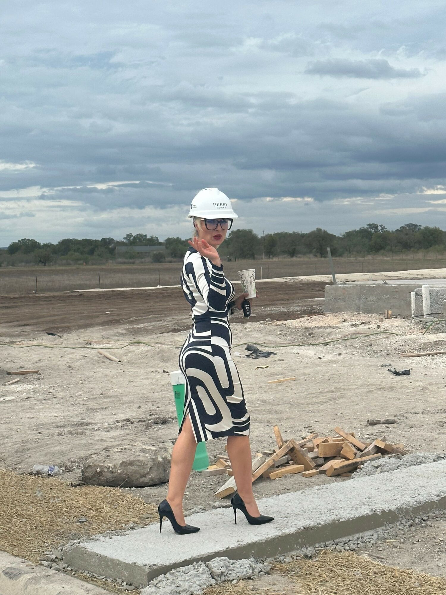 Person in a patterned dress and white hat standing outdoors on a construction site, with cloudy sky and trees in background.