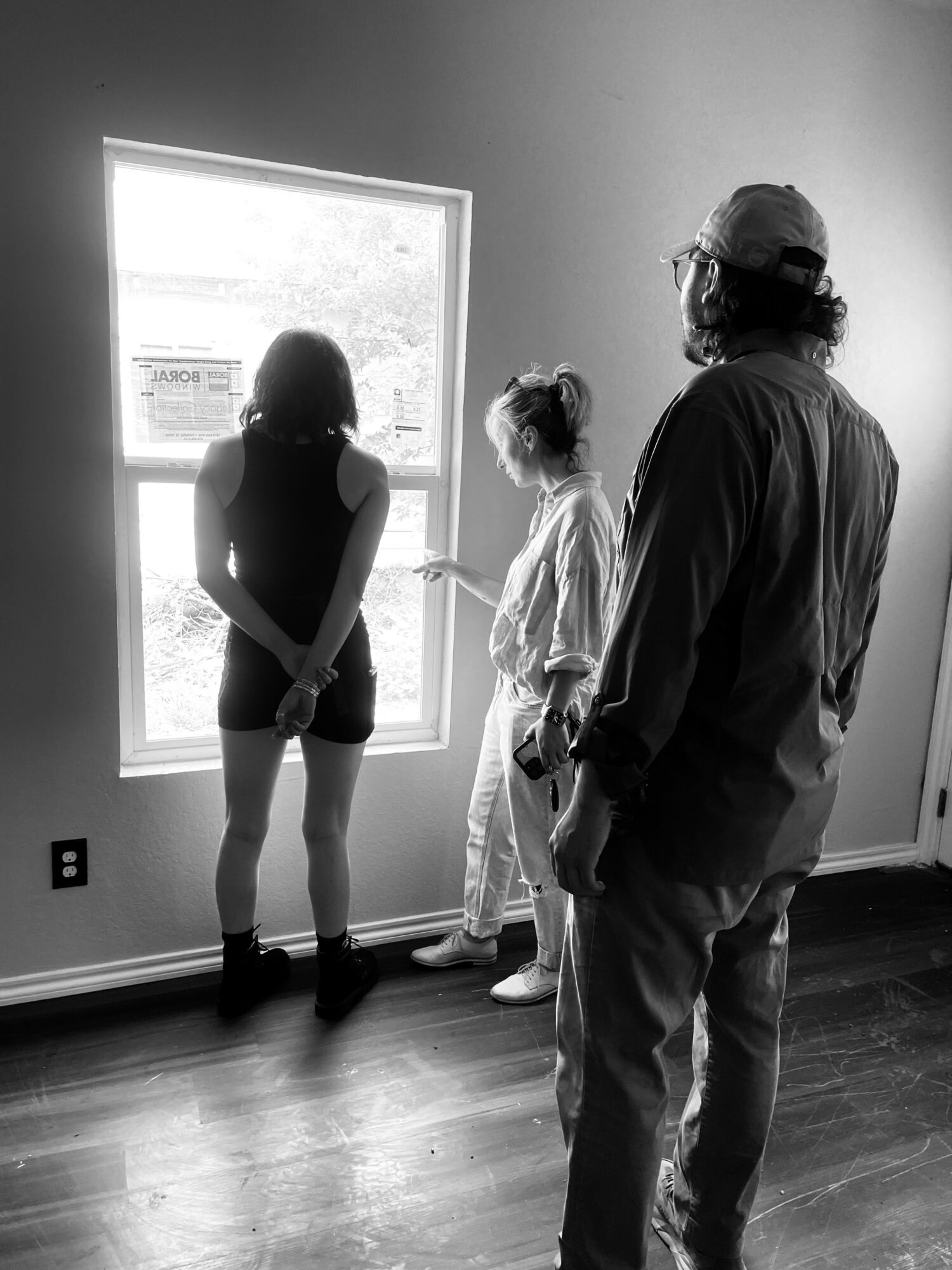 Three people stand near a window, looking outside, with one person facing away and two facing the window, in a room with wooden floor.