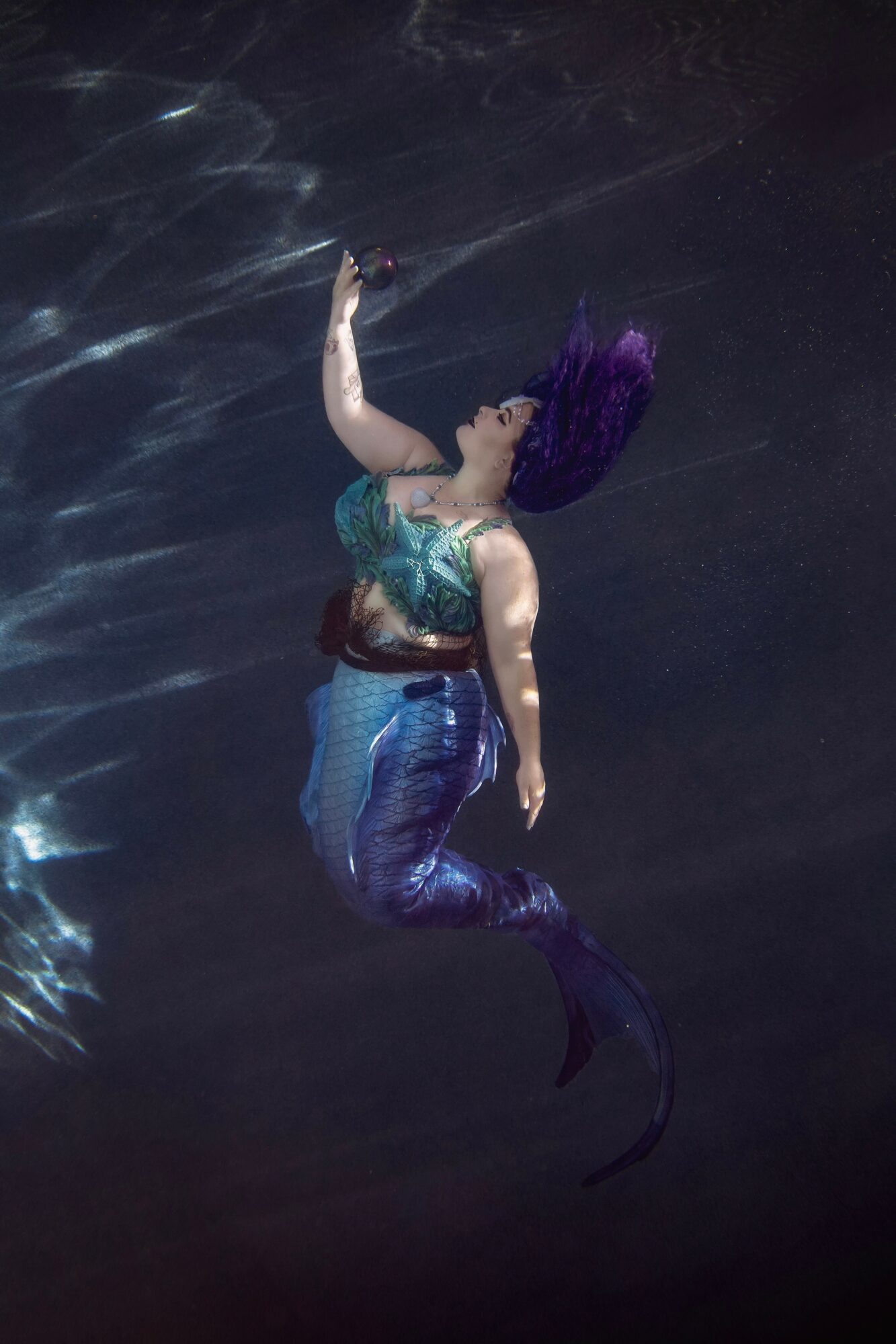 Mermaid with purple hair swimming underwater, reaching upward with one arm, wearing a colorful top and blue tail.