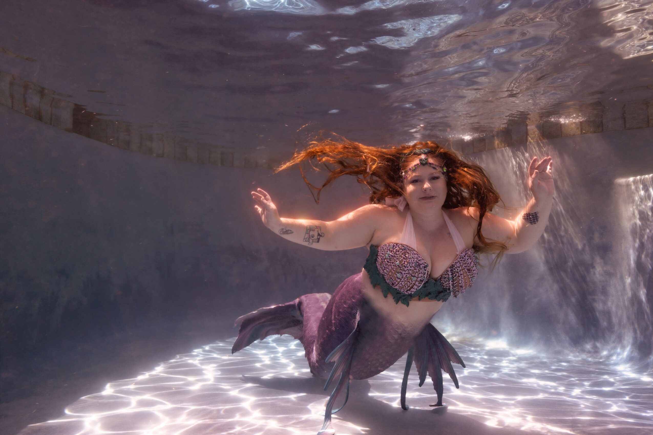 Woman with long red hair swimming underwater, wearing a patterned top and purple skirt, with water flowing around her.