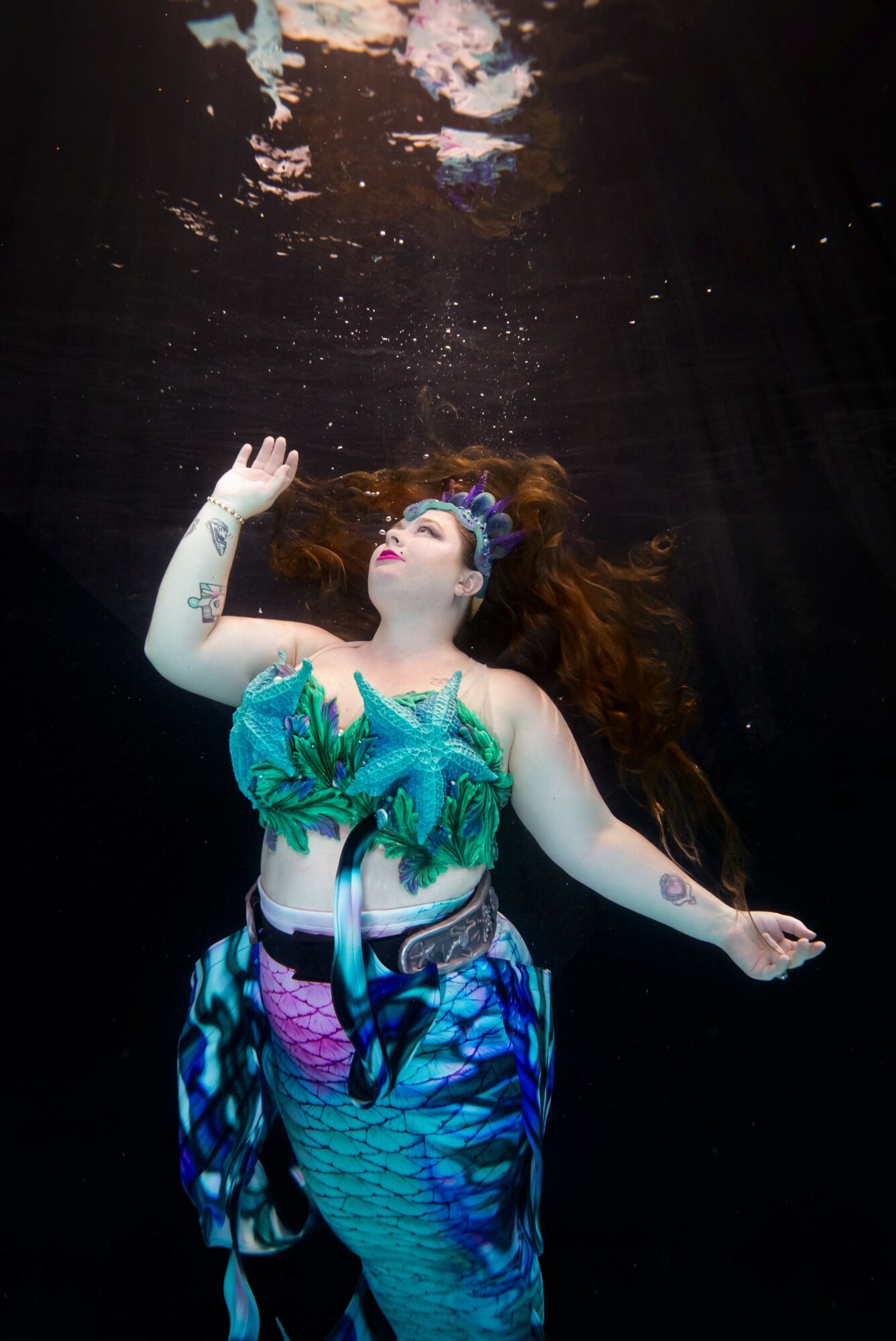 Woman in colorful mermaid costume underwater, hair floating, reaching upward with bubbles above her head.