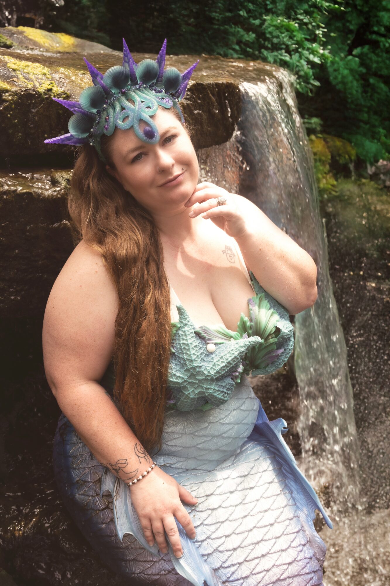 Woman with long hair wearing a mermaid costume and a dragon-themed headpiece, standing near a waterfall in a forest.