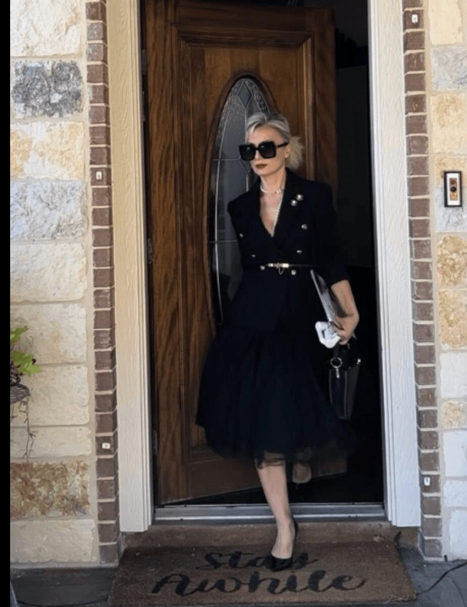 Woman in black dress and sunglasses standing in doorway holding a clutch bag, with a wooden door and brick wall background.