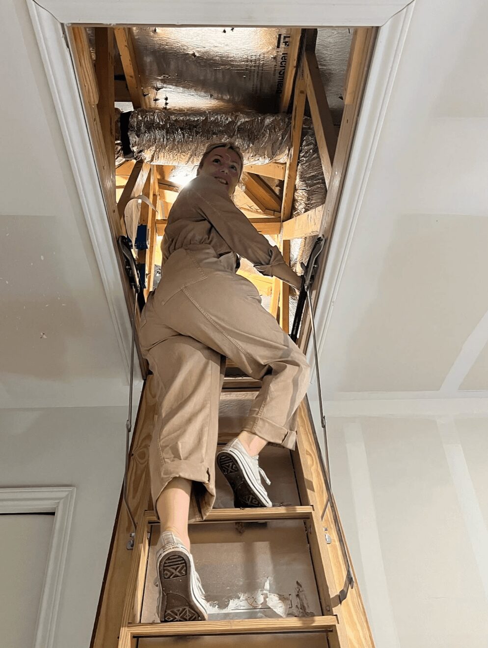 Person climbing a wooden ladder in an attic, looking back over shoulder, with insulation and wooden beams above.