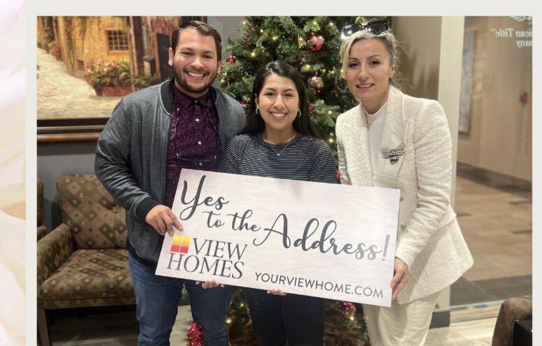 Three people holding a sign in front of a Christmas tree, smiling, indoors, with a couch and painting in background.