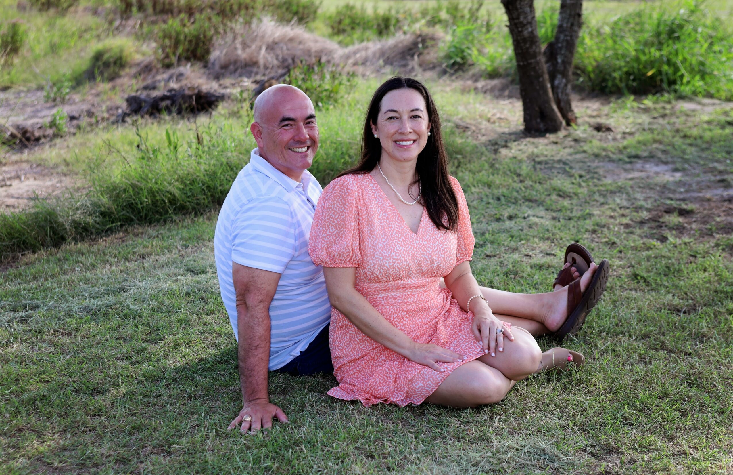A man and woman sitting on grass outdoors, smiling, with trees and greenery in the background.