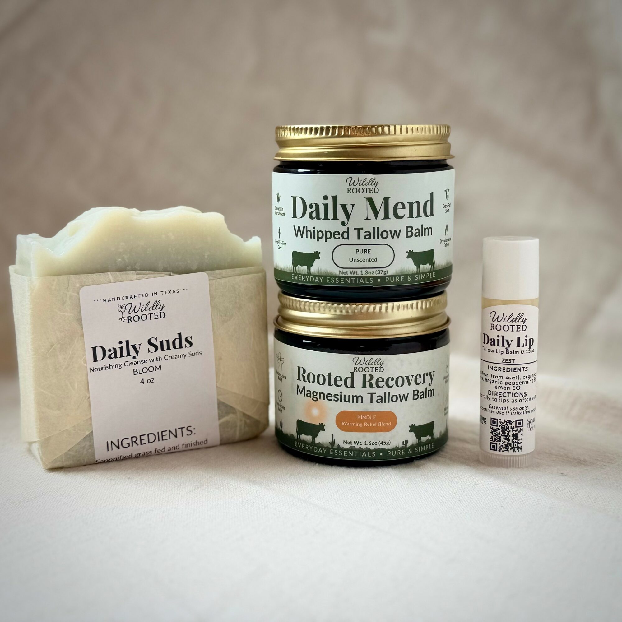 Three skincare products and a soap bar on a surface, with a neutral background. Two jars labeled 'Daily Mend' and 'Rooted Recovery', and a small tube.