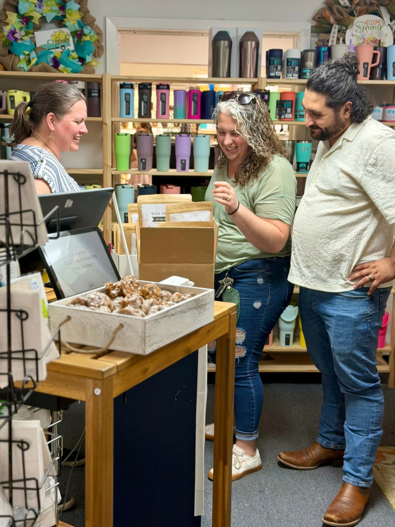 Three people stand at a store counter, smiling and talking, with shelves of colorful items behind them.