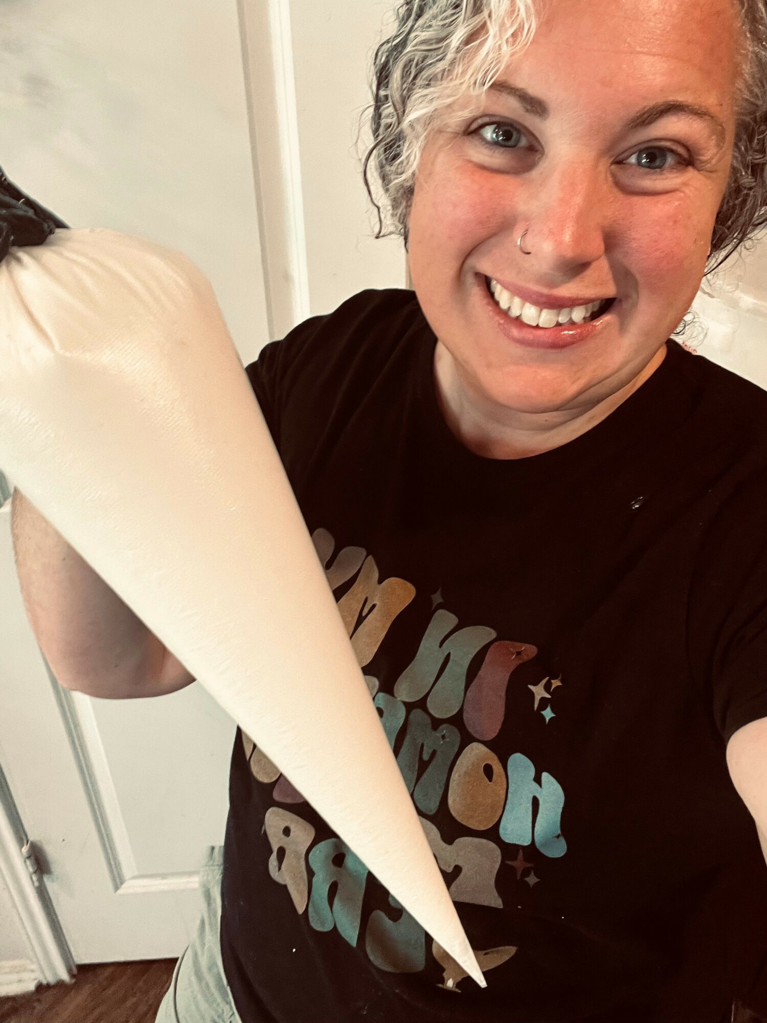 Person smiling and holding a large piping bag filled with cream or frosting, standing indoors.