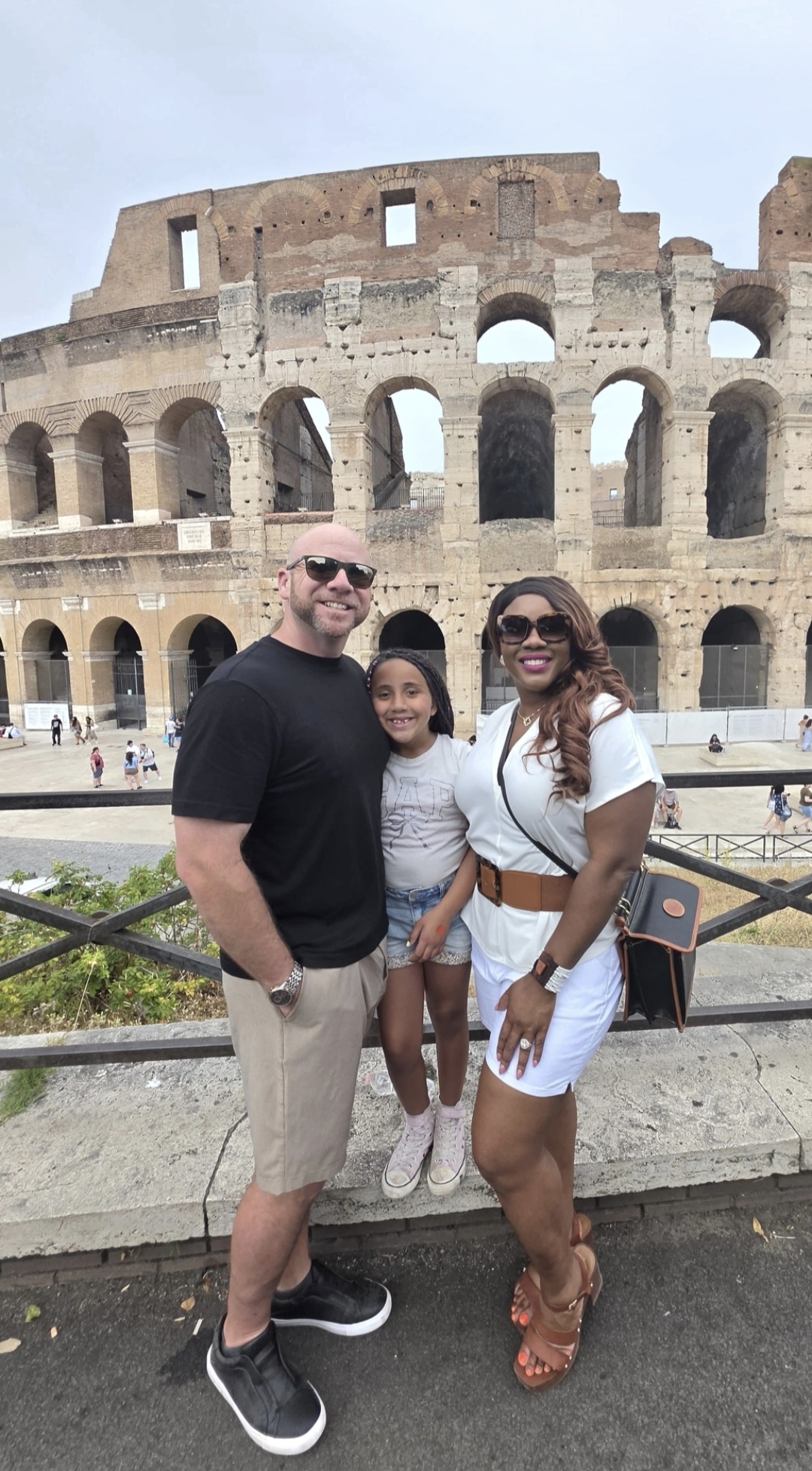 Three people standing in front of the Colosseum, smiling, with a railing and greenery in the foreground.