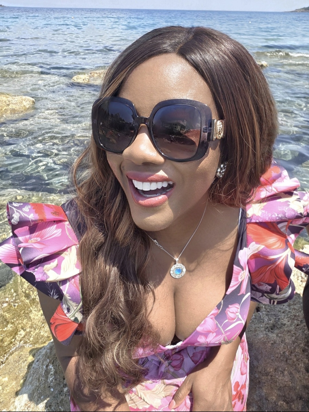 Woman with long hair wearing sunglasses and a floral dress at the beach, smiling, with water and rocks in background.