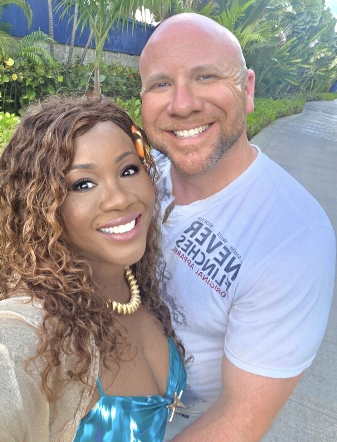 A smiling couple outdoors, woman with curly hair and necklace, man with a shaved head, both in casual clothing.