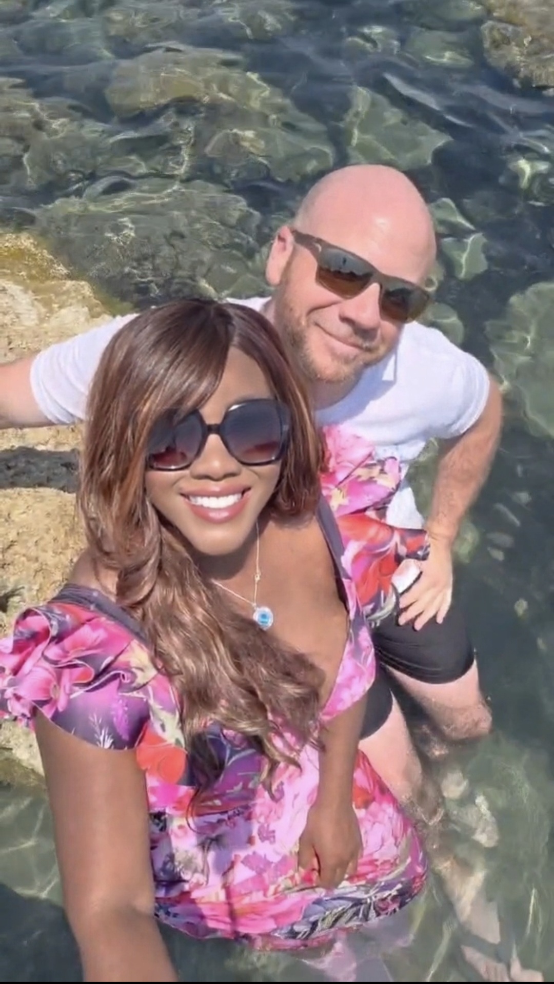 Two people standing in water near rocks, smiling, wearing sunglasses, one in a pink floral top, the other in a white shirt.