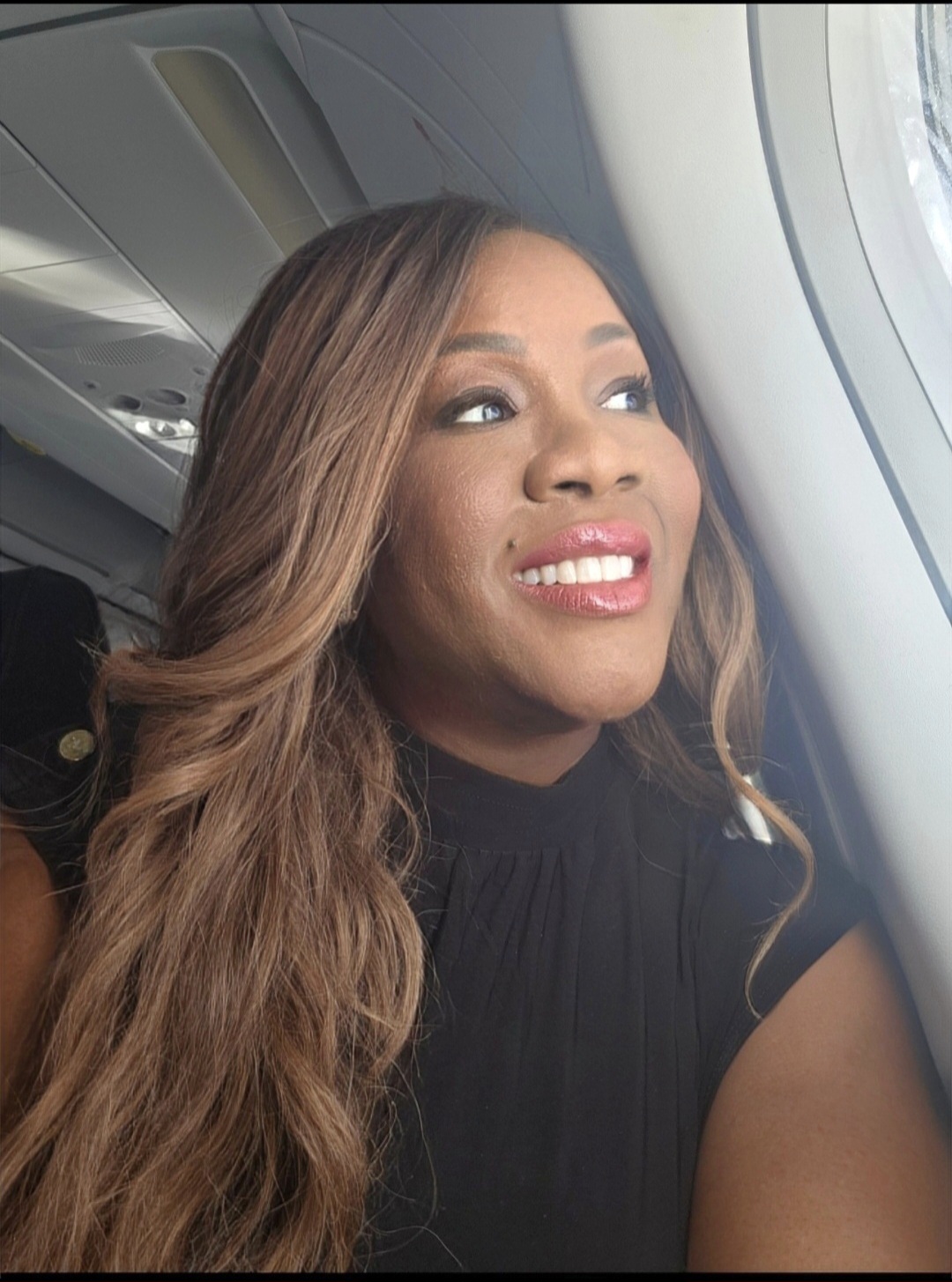 Woman looking out airplane window with a smile, long wavy hair, and black top.