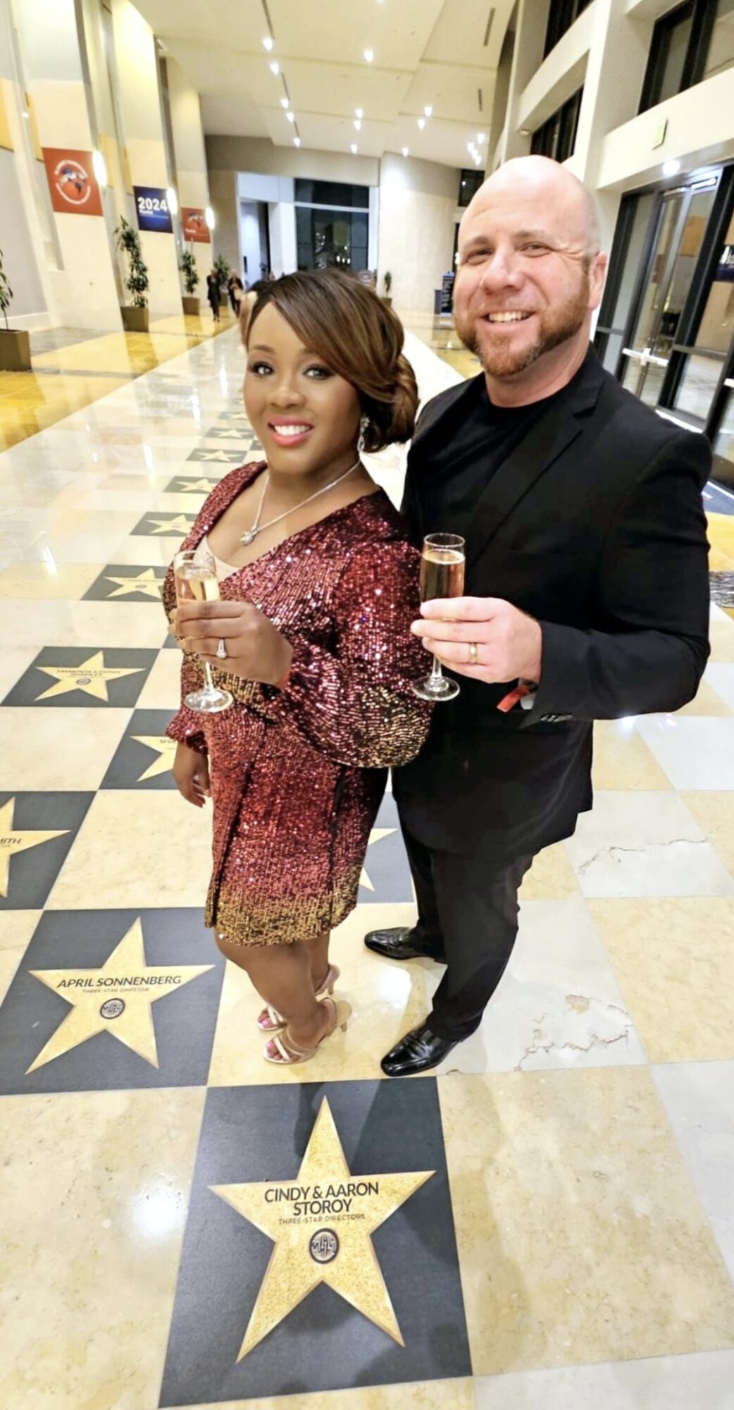 Two people in formal attire holding glasses in a hallway with star-shaped floor markers, smiling at the camera.