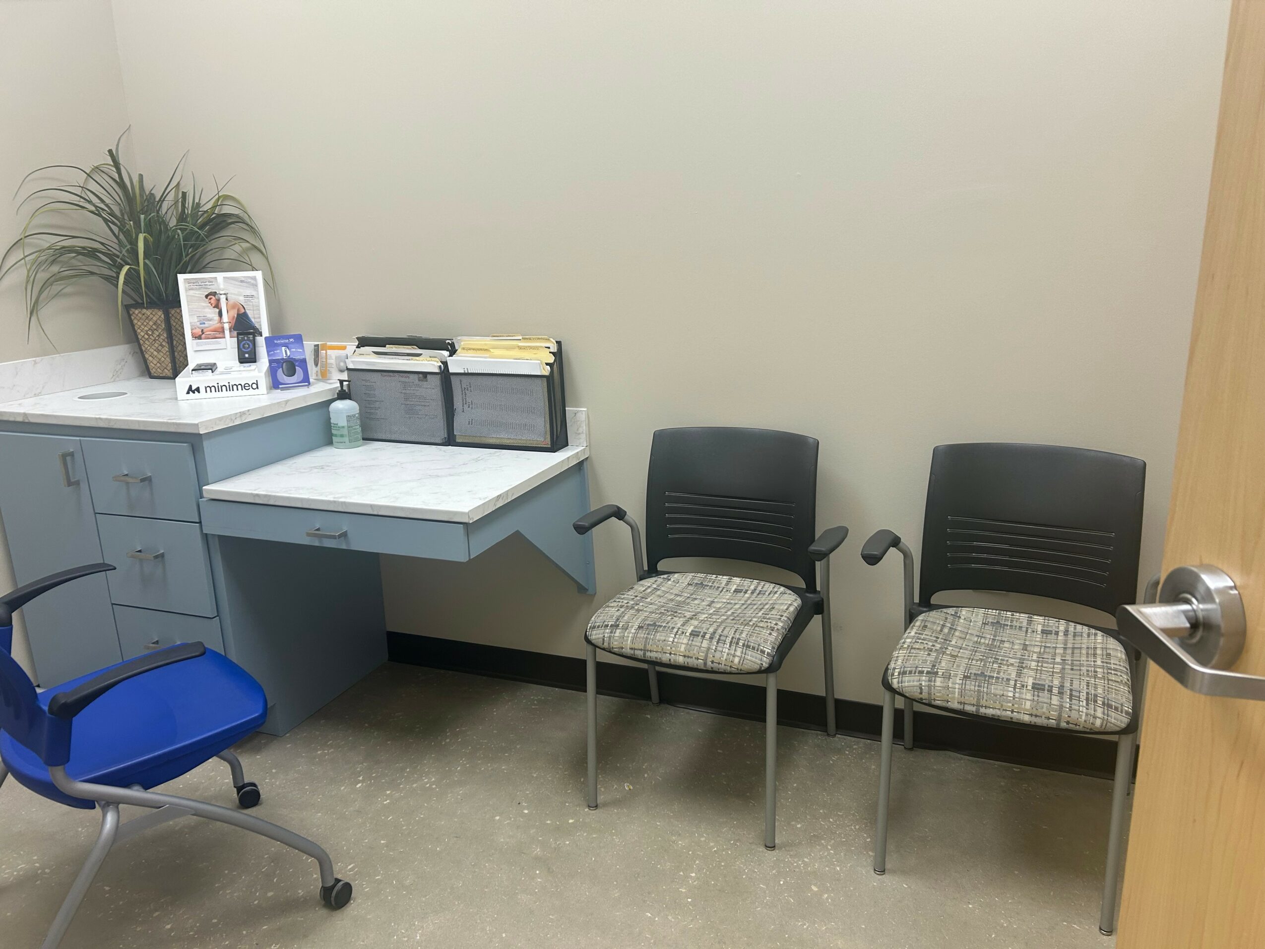Office waiting area with two black chairs, a blue chair, a desk with files, and a potted plant on a cabinet.