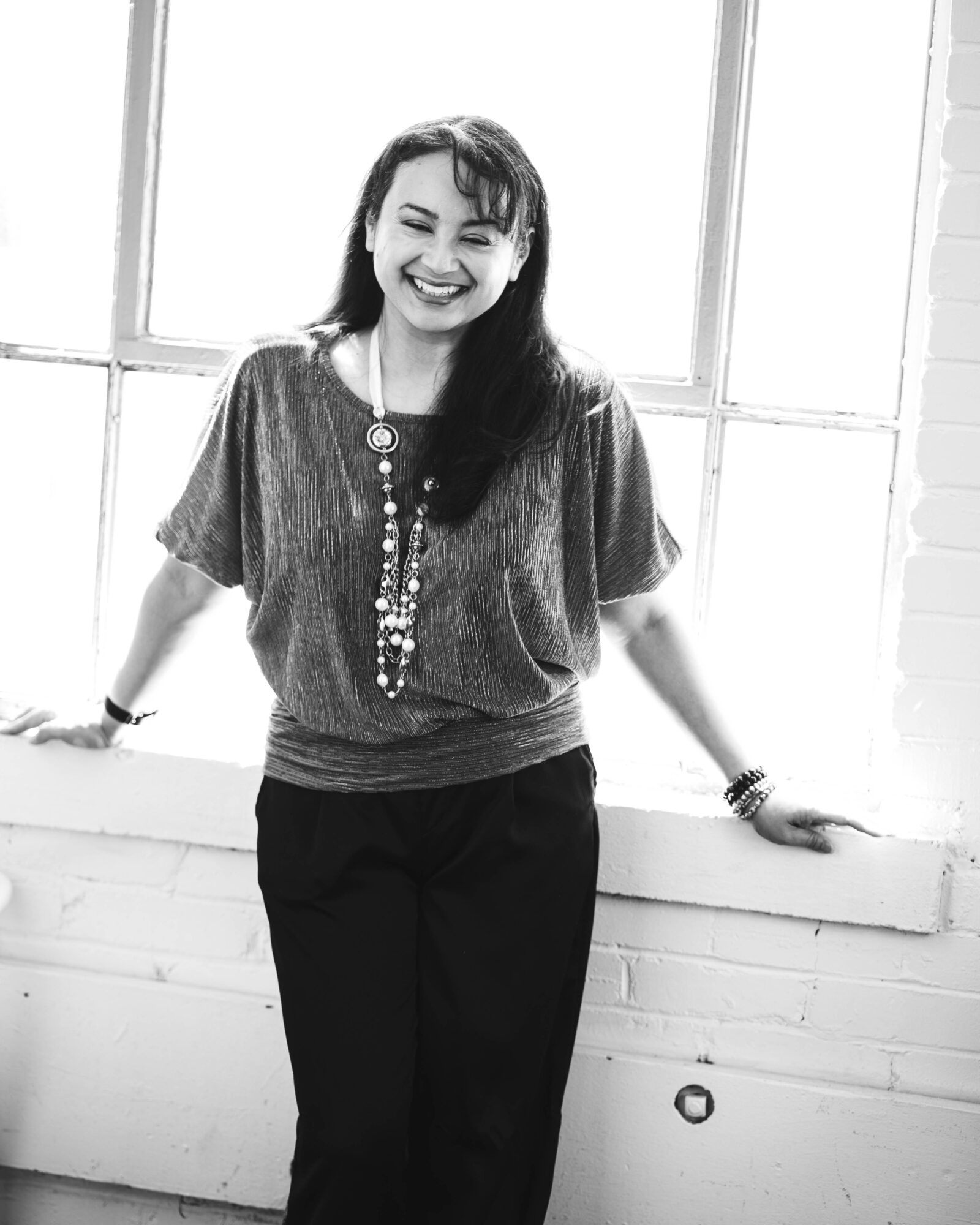 Woman smiling, standing by a window with brick wall, wearing a top and necklace, in black and white.