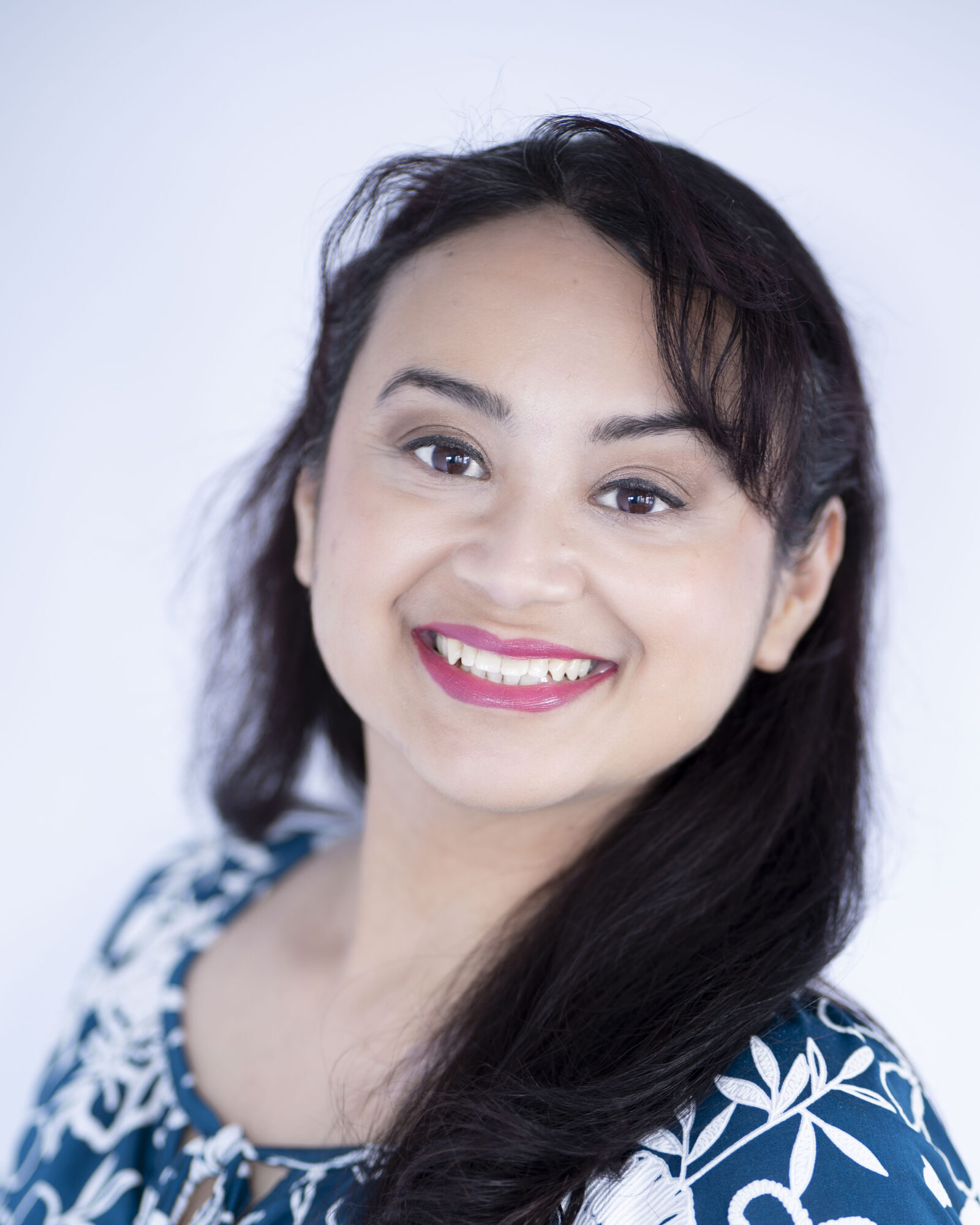 Smiling woman with long dark hair, wearing a blue and white patterned top, against a light background.