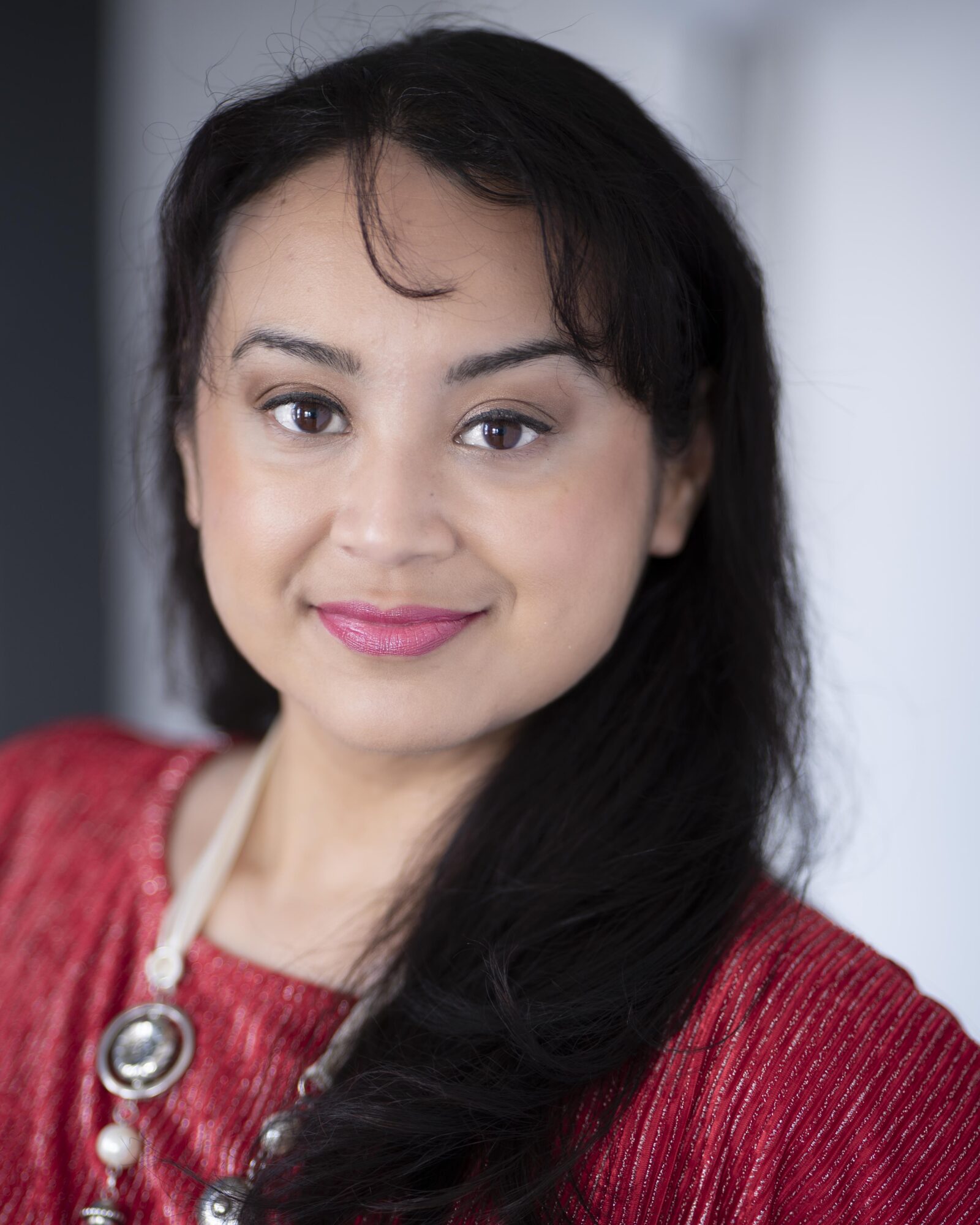 Portrait of a woman with dark hair, wearing a red top and a necklace, smiling softly.