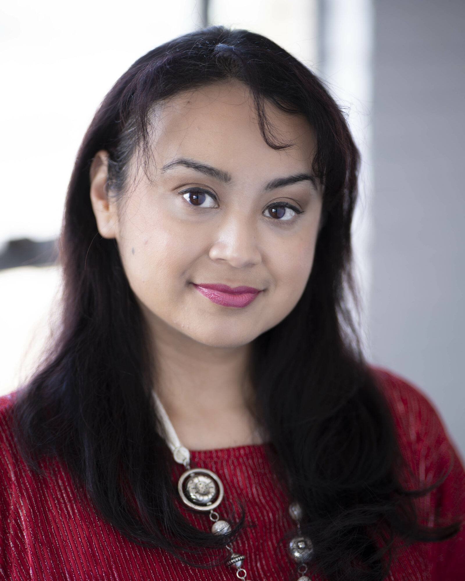 Young woman with long dark hair, wearing a red top and necklace, smiling at the camera.