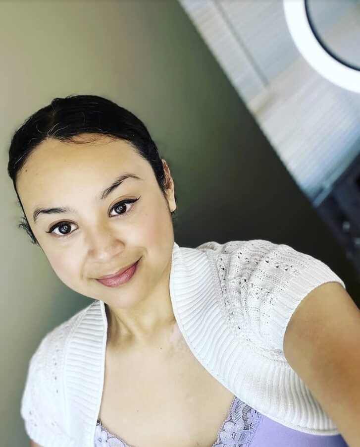 Young woman with dark hair in a bun, smiling, wearing a white top, in front of a green wall and a washing machine.