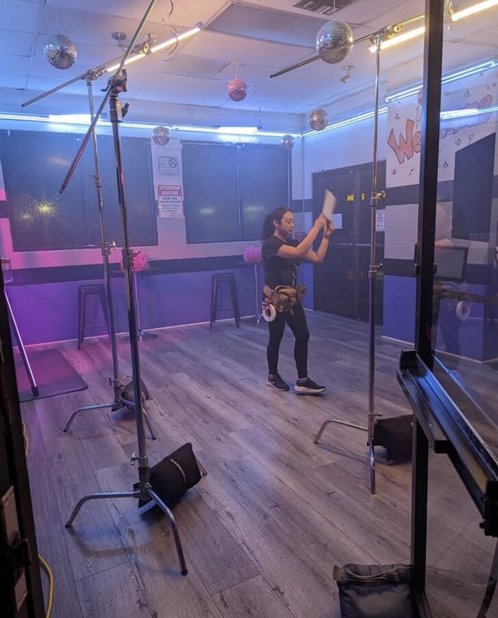 Person standing in a dance studio, holding a paper, with colorful lighting and mirrors on the walls.