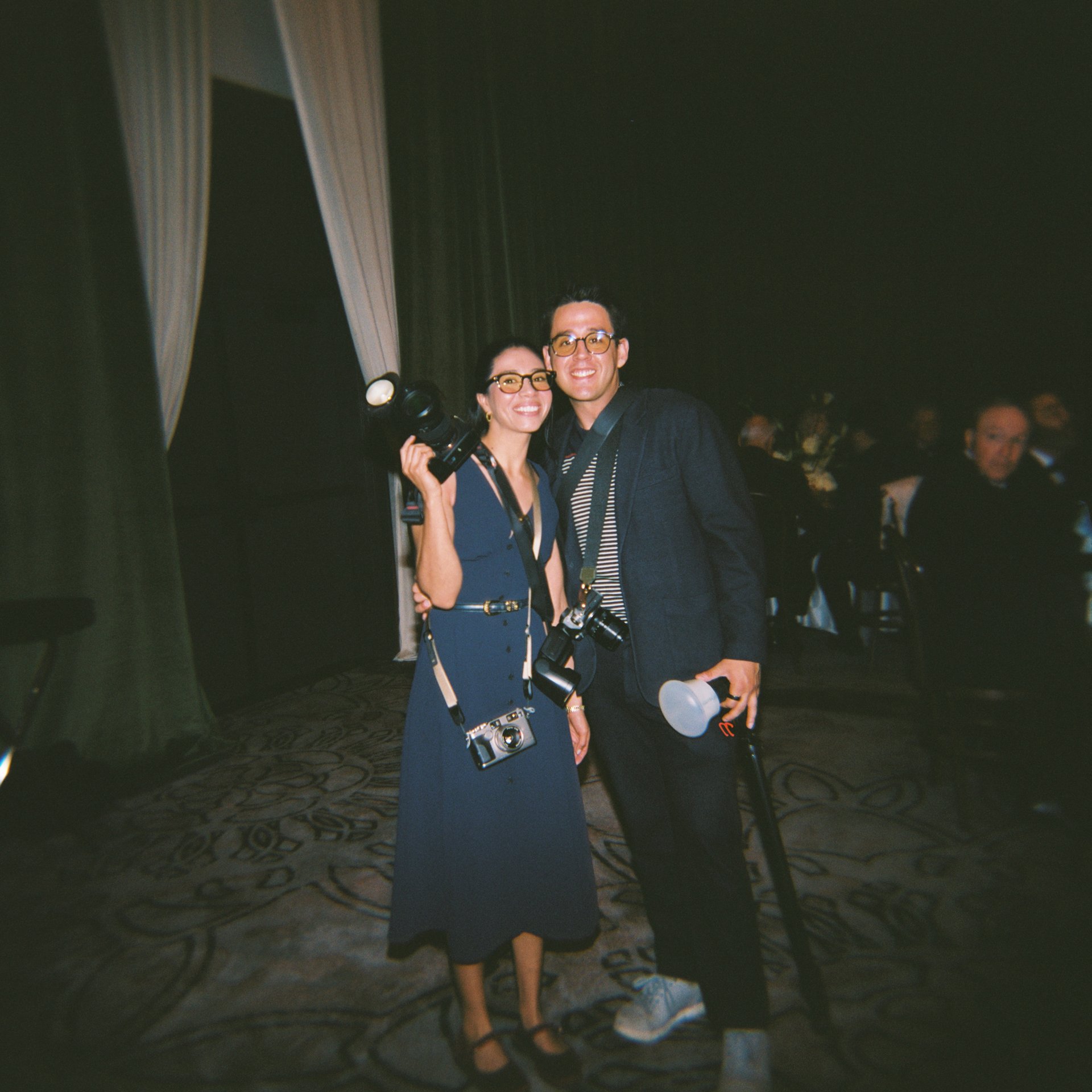 Two people standing together in a dimly lit room, smiling, with tables and other guests in the background.