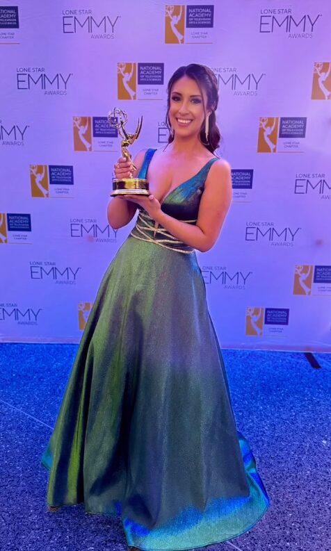 Woman in a green and blue gown holding an Emmy award, standing in front of a backdrop with Emmy logos.