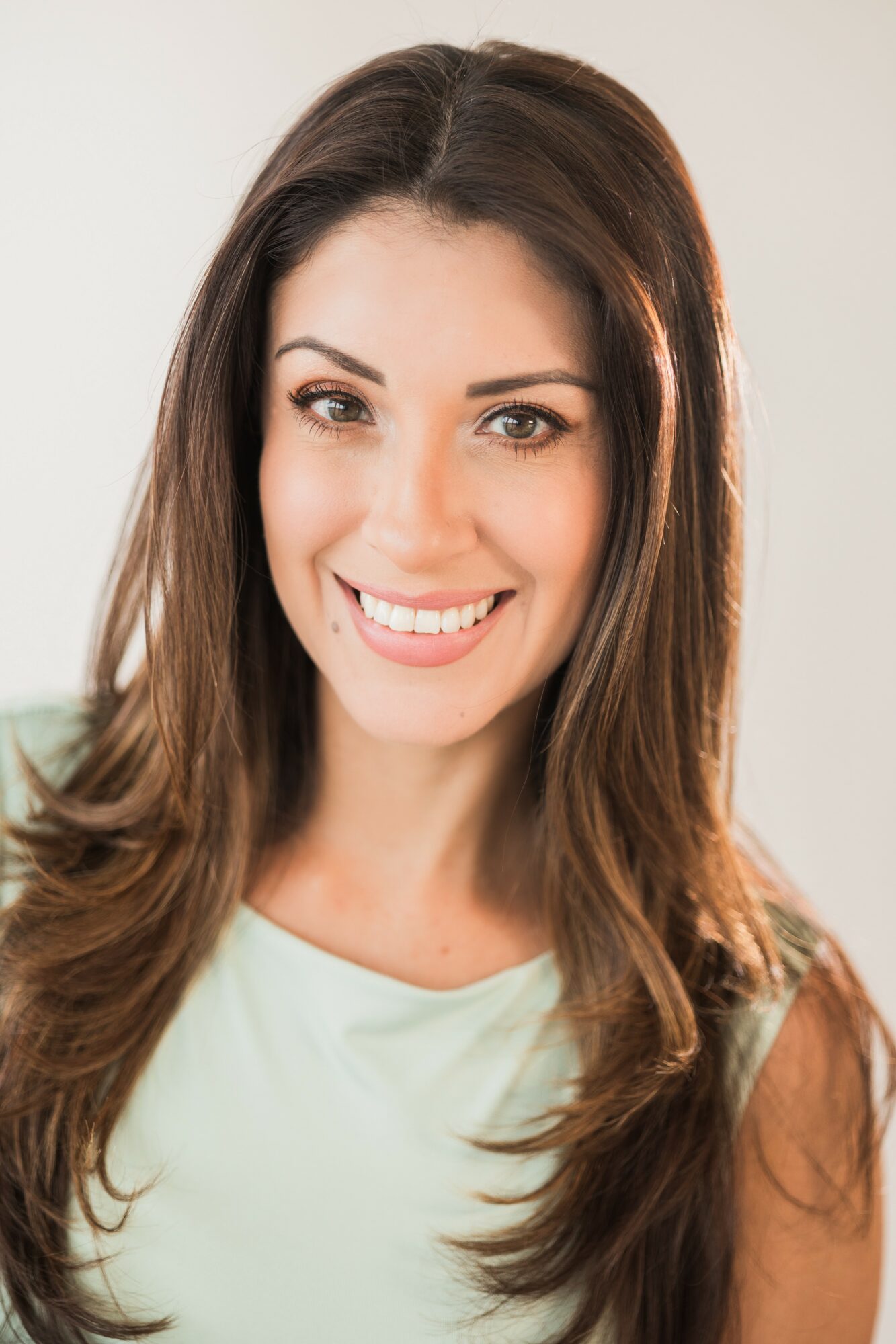 Smiling woman with long brown hair and light green top, looking at camera, neutral background.