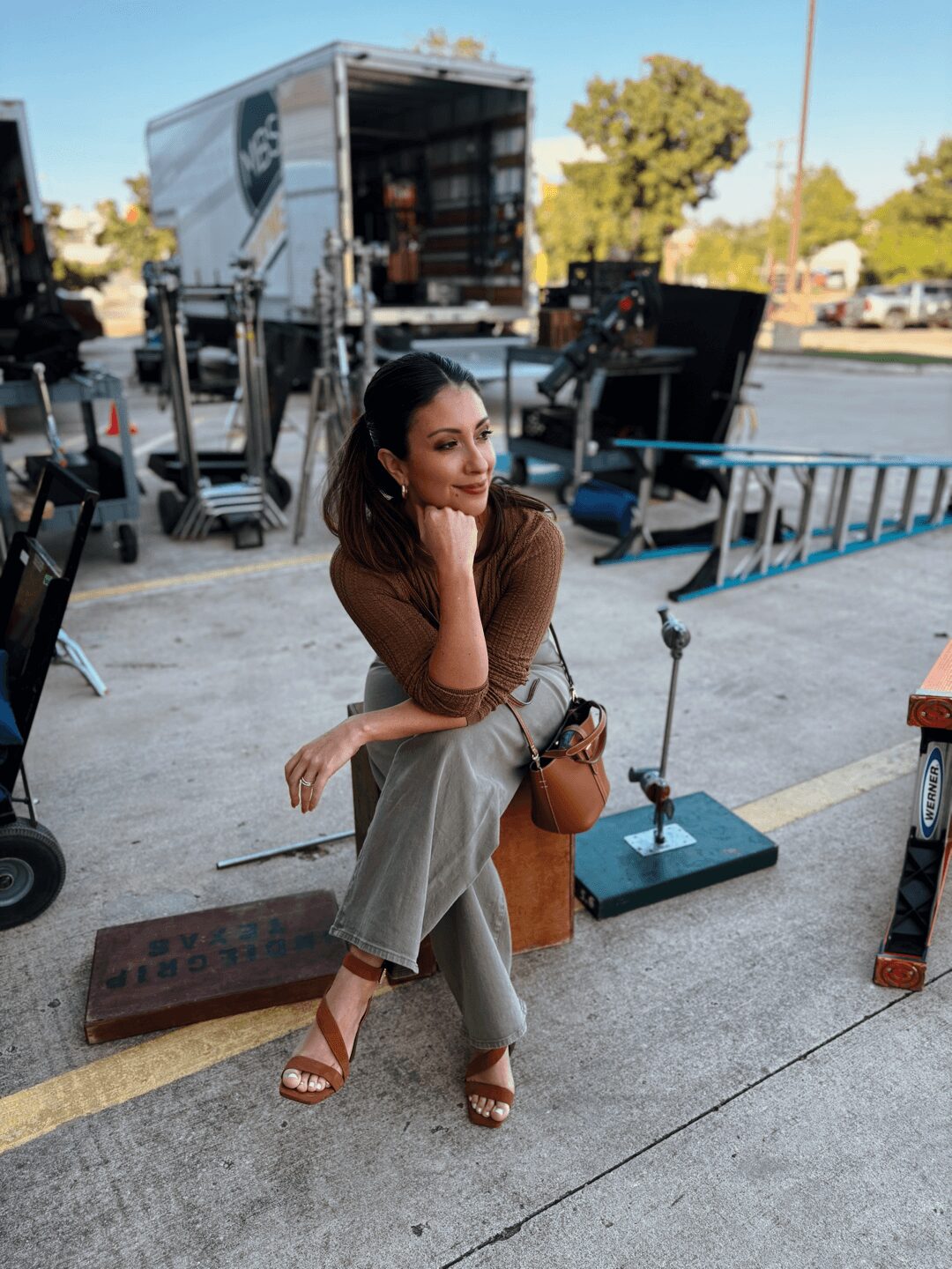 Woman sitting on a box outdoors, surrounded by equipment, with trees and vehicles in the background.
