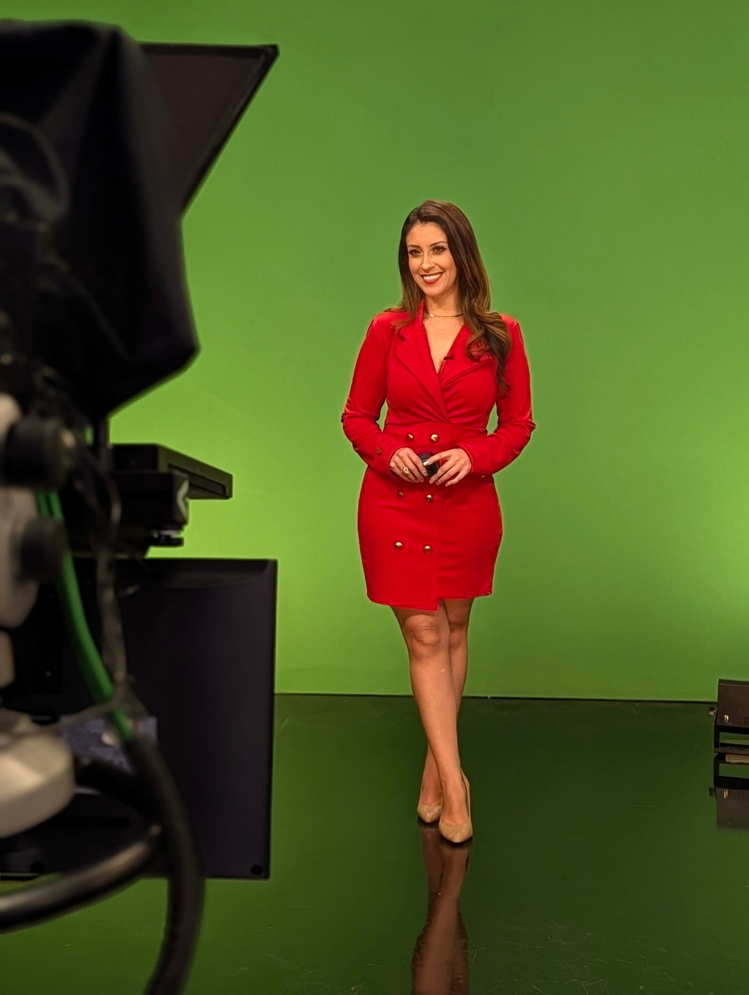 Woman in red dress standing in front of green screen, smiling, with camera equipment visible, studio setting.