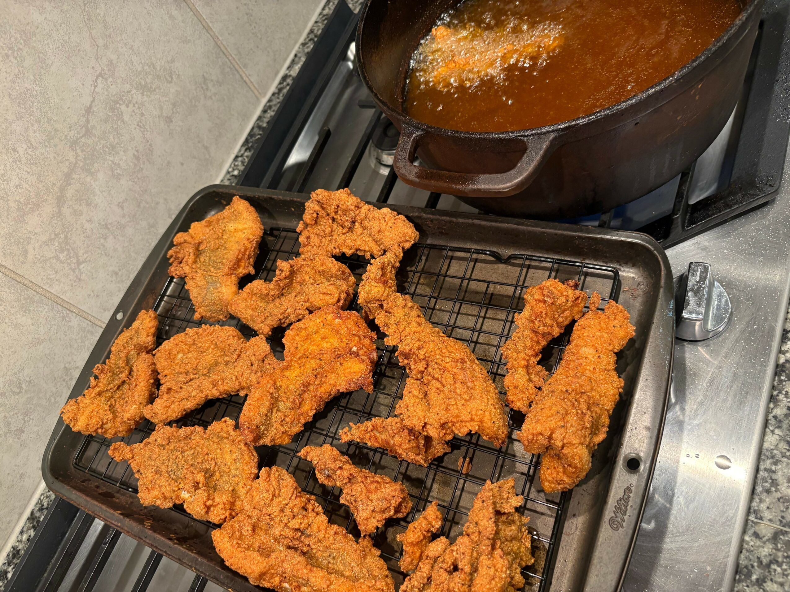 Fried chicken pieces on a wire rack, with a pot of hot oil on a stove in the background.