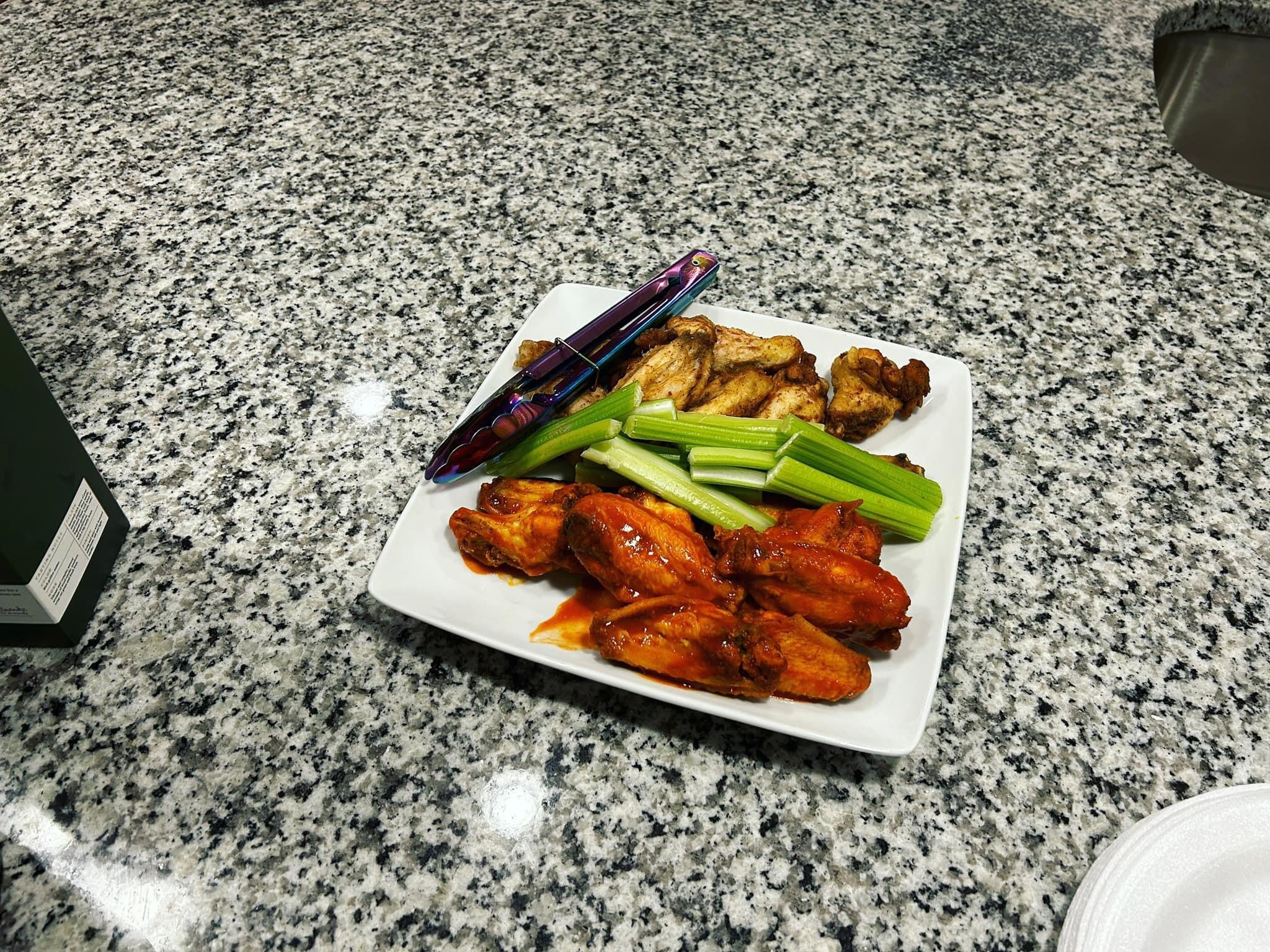 Square white plate with chicken wings, celery sticks, and a dipping sauce on a granite countertop.