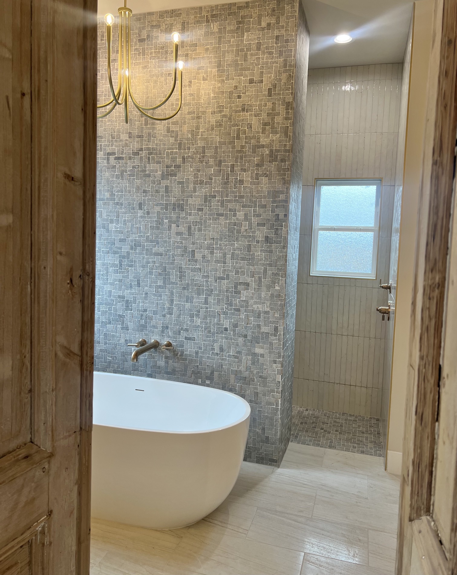 Bathroom with a freestanding bathtub, textured wall, window, and chandelier, viewed through a wooden doorway.