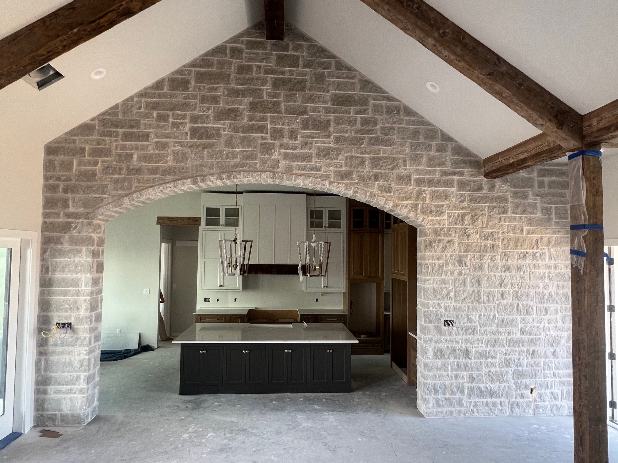 Interior view of a room with a brick archway opening to a kitchen area, exposed wooden beams, and white walls.