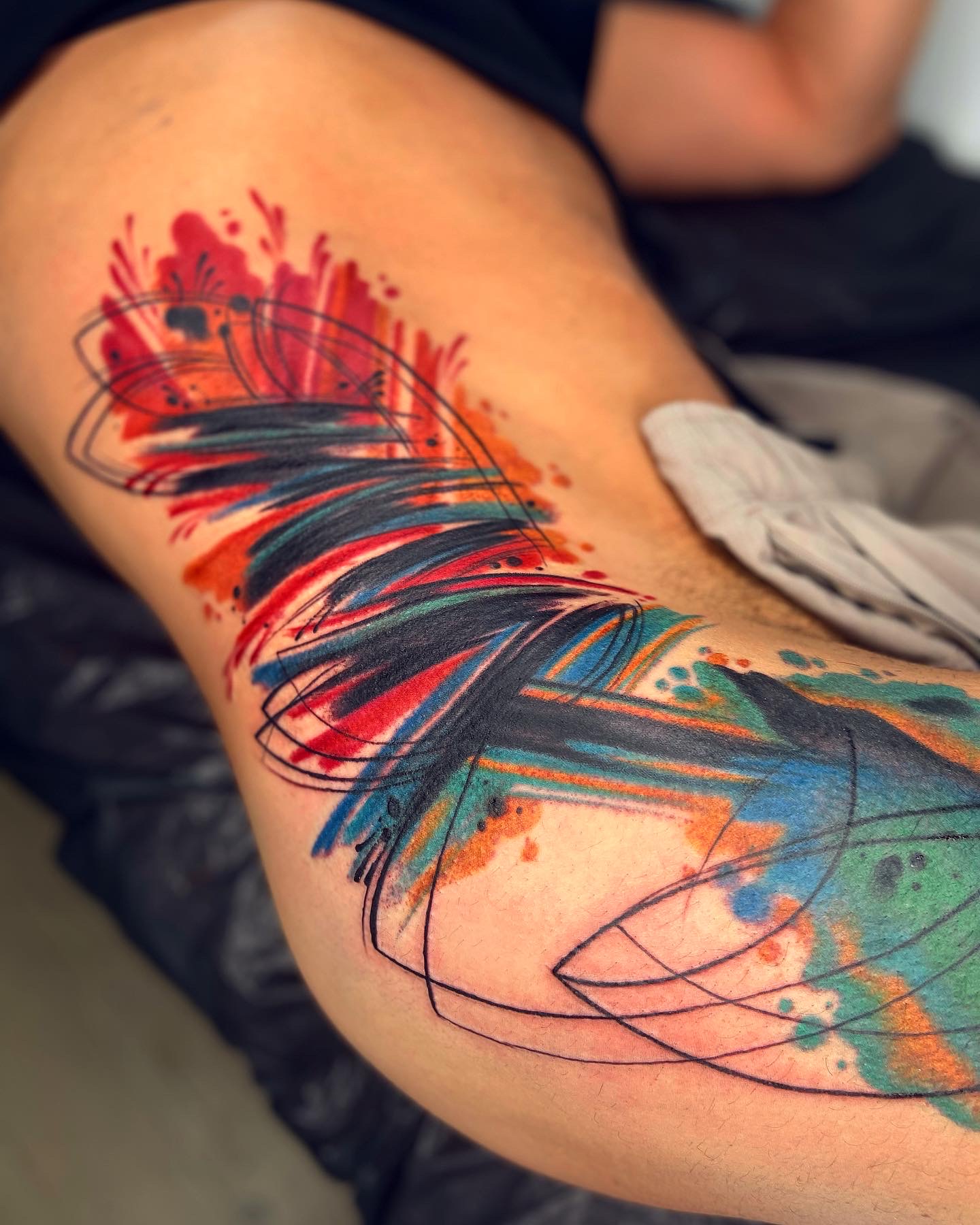 Tattoo of colorful abstract lines and splashes on a person's thigh, with a cloth nearby.
