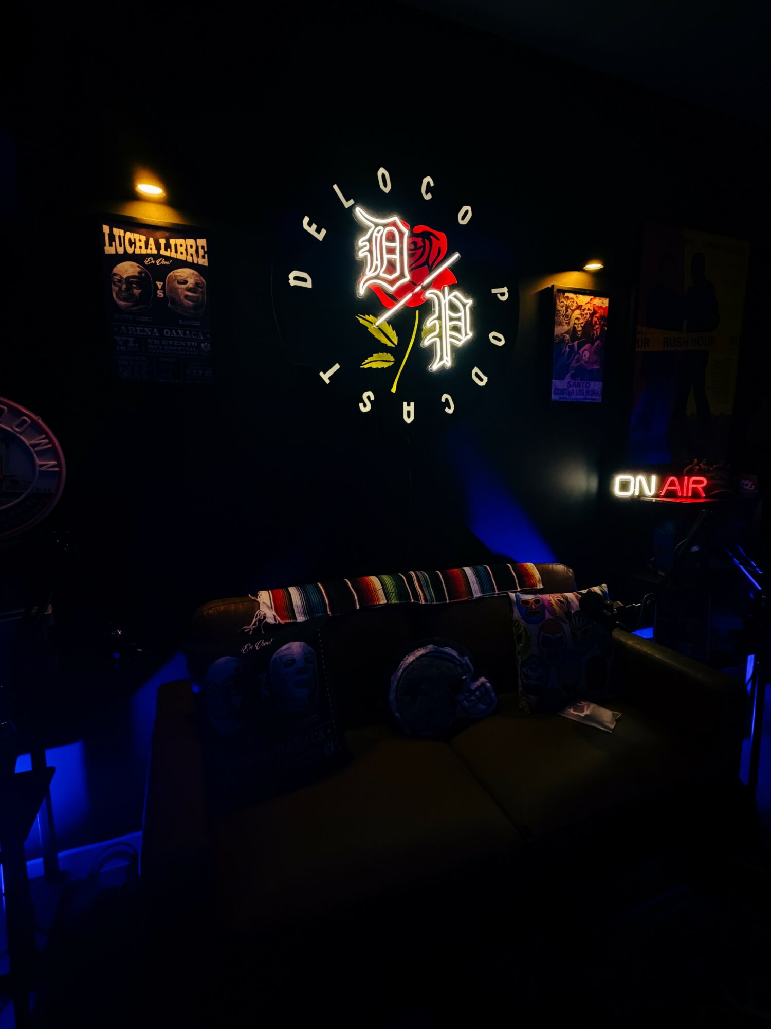Dark room with illuminated Detroit logo, posters, and an ON AIR sign, with a couch and colorful blanket.