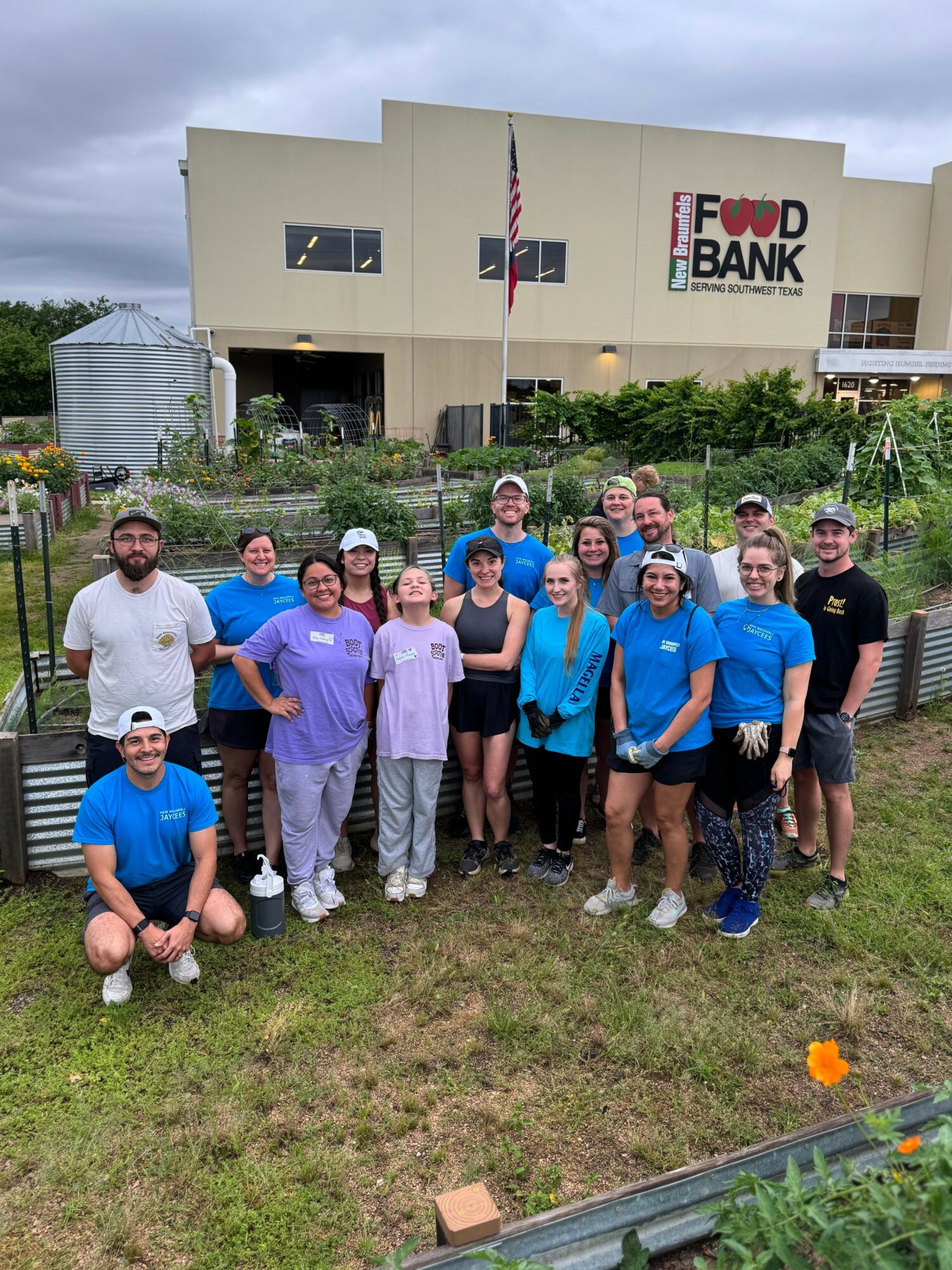 Group of people standing outdoors in front of a building with a sign that reads 'Food Bank,' some wearing purple and blue shirts, on cloudy day.