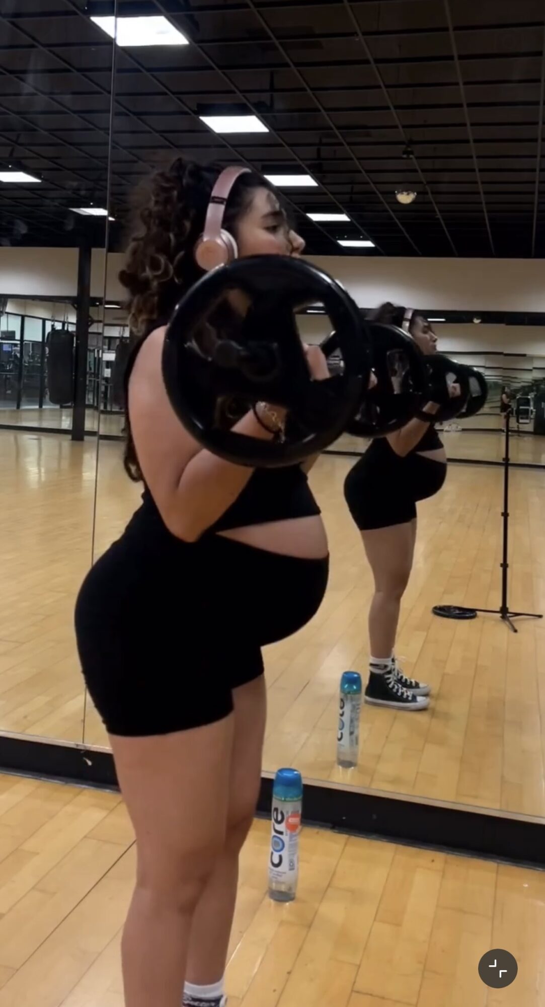 Woman in gym holding a weight plate, mirror reflection shows her workout attire and gym environment.