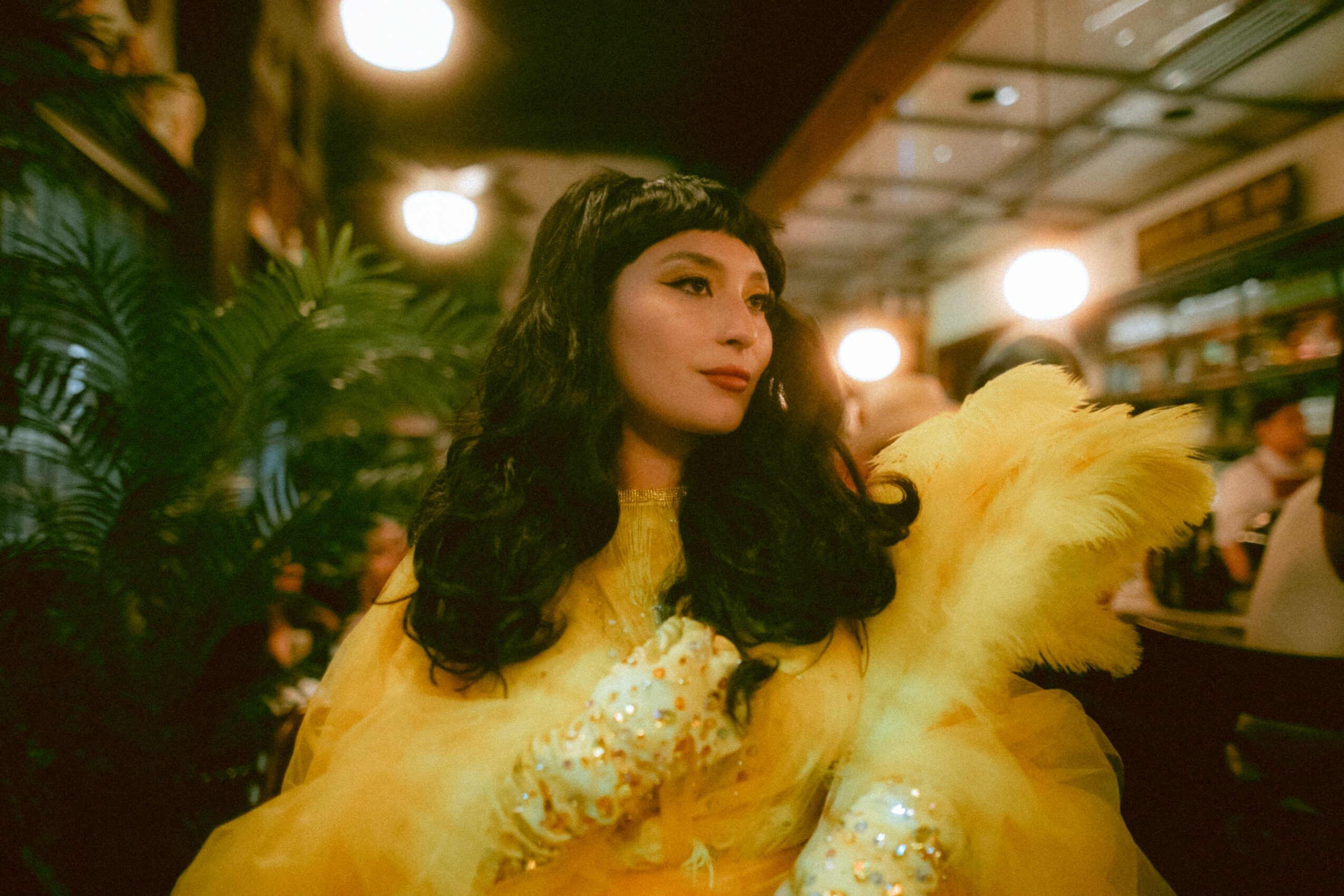 Woman with dark wavy hair wearing a yellow dress and feathered shawl in a lively indoor setting.