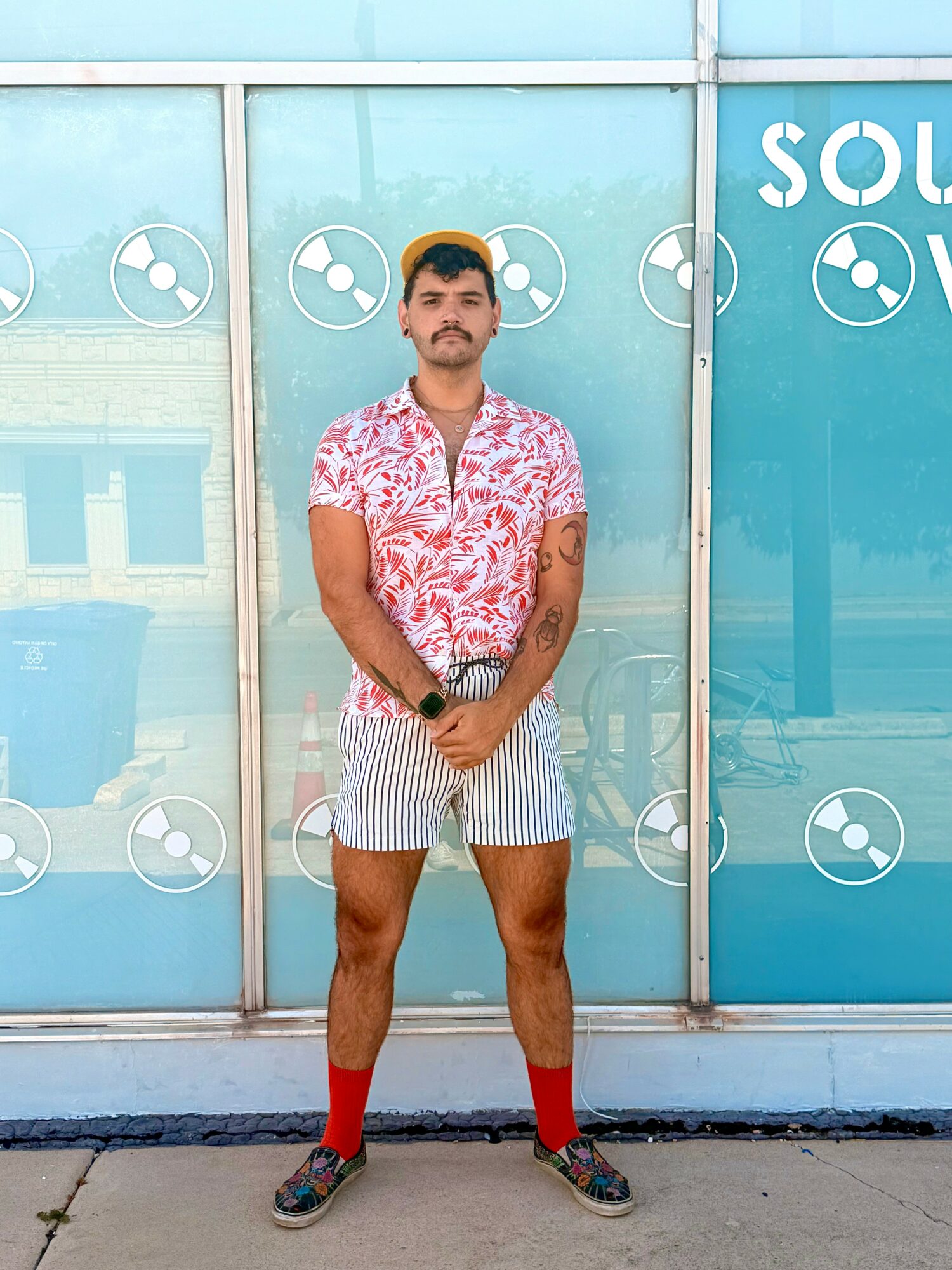 Man standing outdoors in front of a glass wall with logos, wearing a floral shirt, striped shorts, and colorful shoes.
