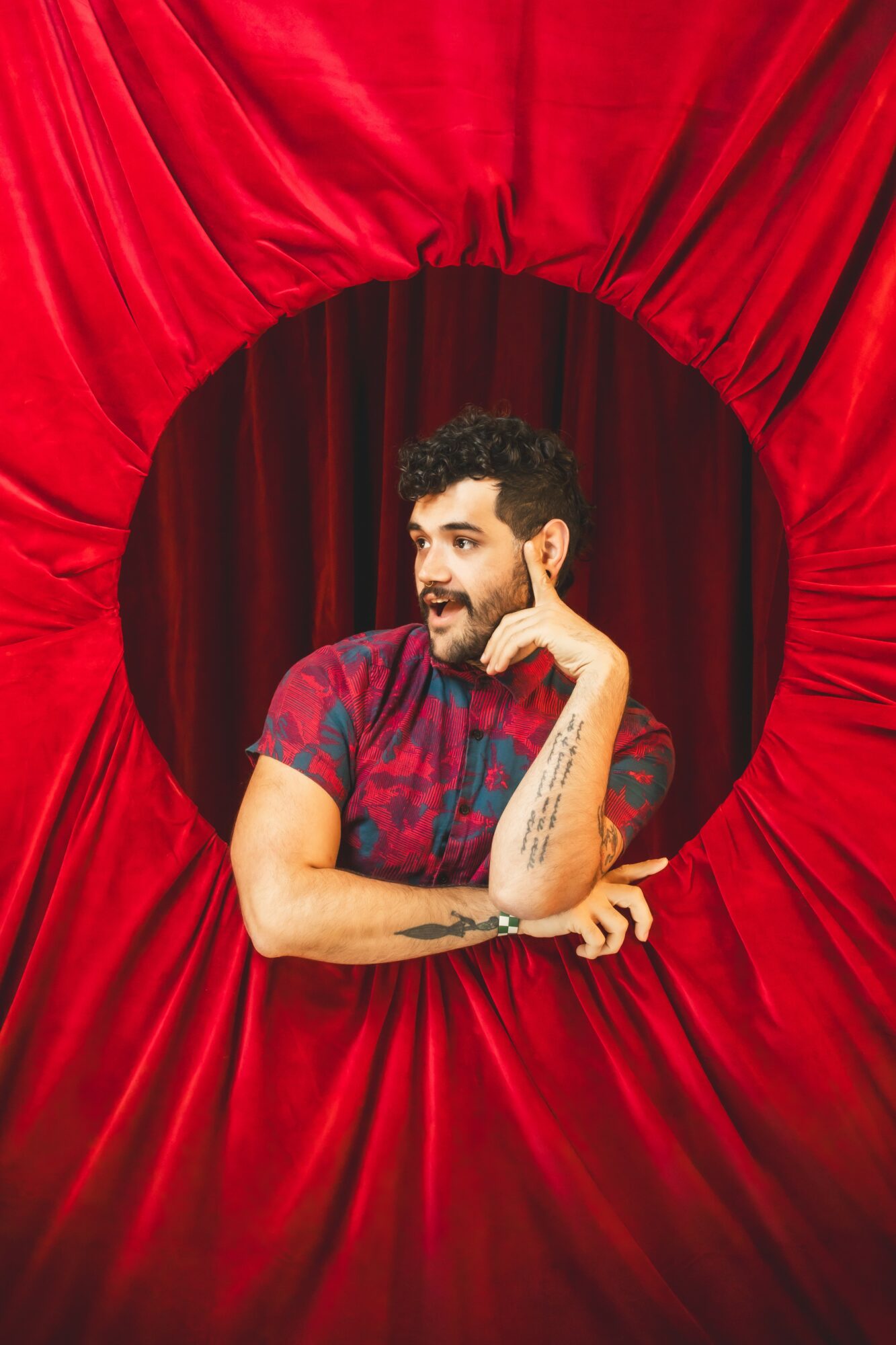 Person with curly hair and beard, wearing a colorful shirt, smiling, resting chin on hand, behind red curtain opening.